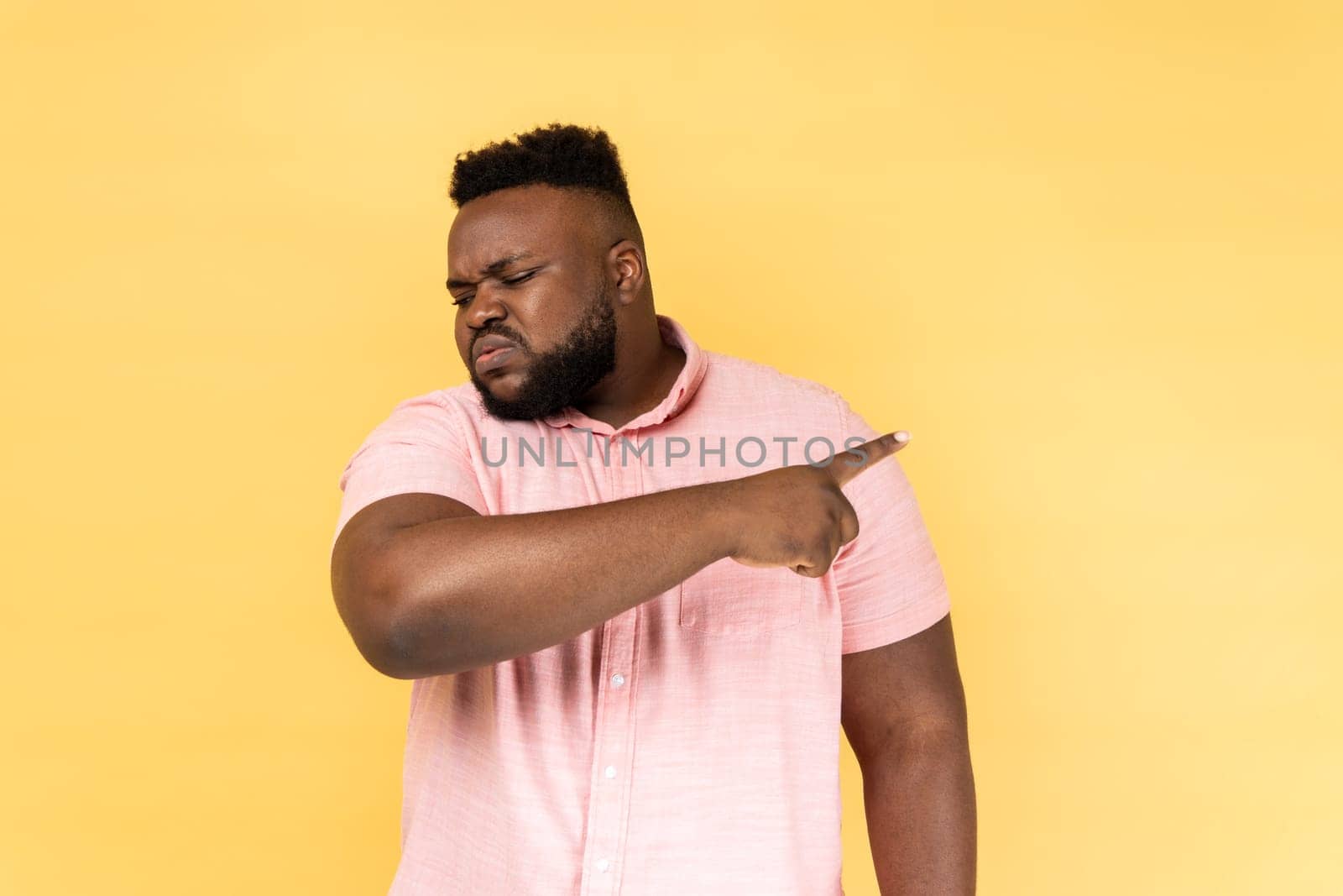Portrait of man wearing pink shirt pointing finger aside, ordering get out and looking resentful, boss dismissing from work, showing exit. Indoor studio shot isolated on yellow background.