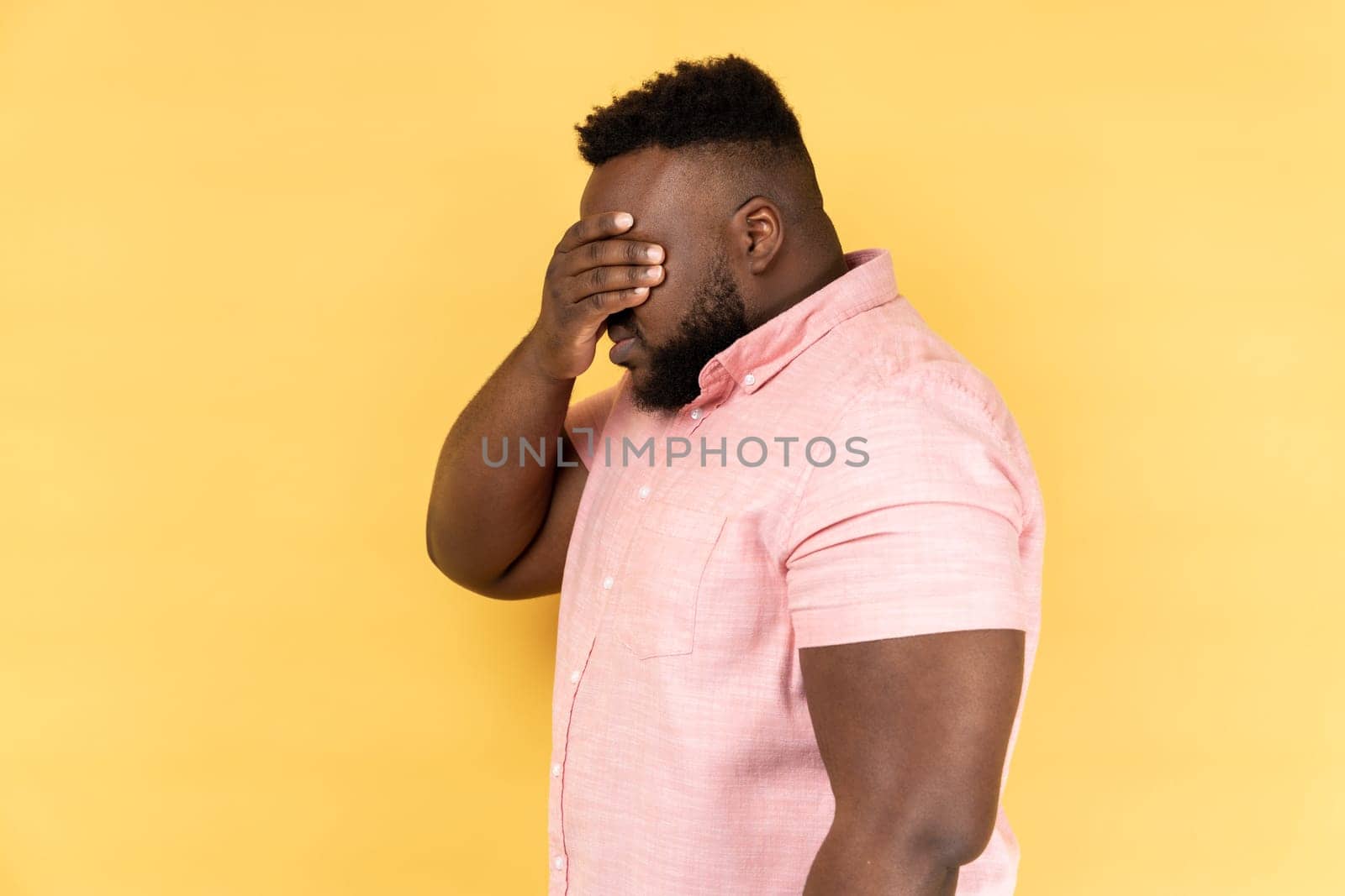 Side view of man wearing pink shirt closing eyes with hand, dont want to see that, ignoring problems, hiding from stressful situations. Indoor studio shot isolated on yellow background.