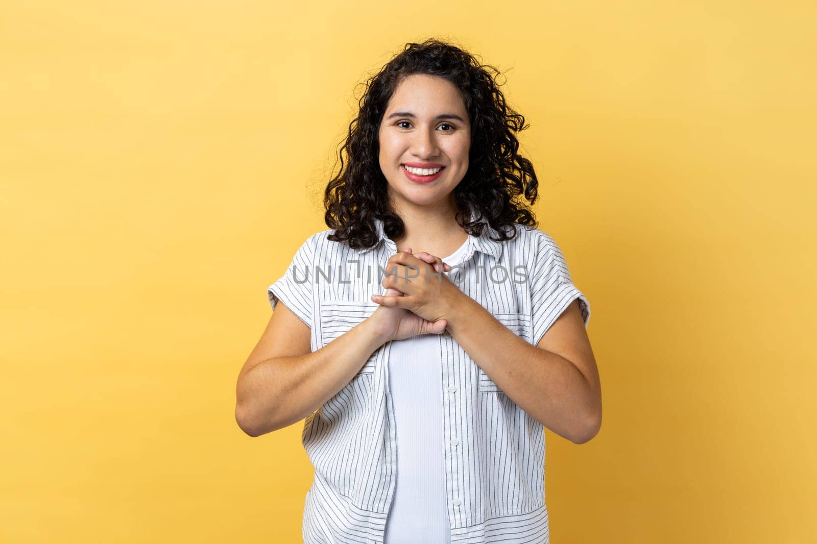 Portrait of positive optimistic friendly woman with dark wavy hair standing looking at camera, keeps hands together, expressing happiness. Indoor studio shot isolated on yellow background.