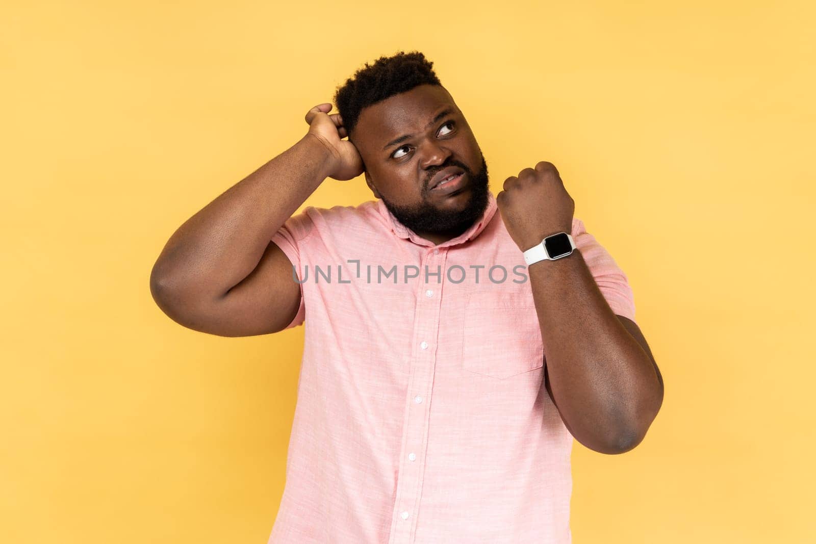 Portrait of puzzled confused man wearing pink shirt showing his smart watch and scratching his head, looking away with with thoughtful expression. Indoor studio shot isolated on yellow background.