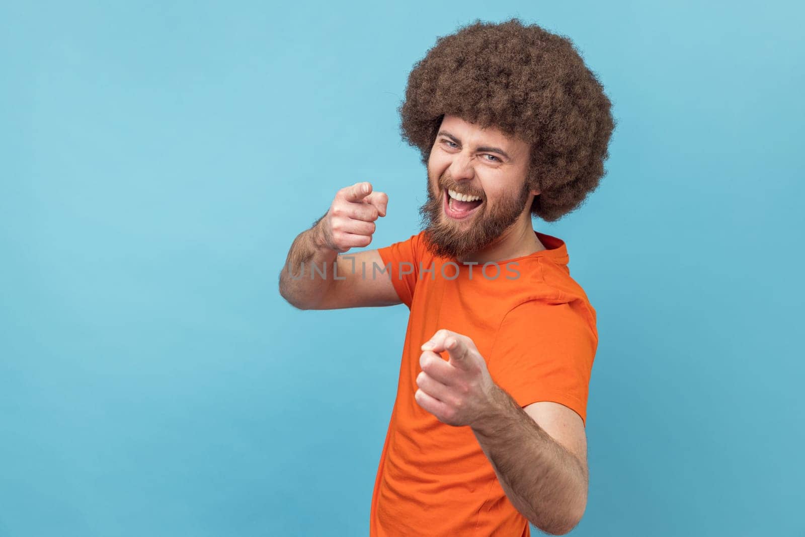 Portrait of man with Afro hairstyle wearing orange T-shirt pointing to camera, smiling, making happy choice, we need you concept. Indoor studio shot isolated on blue background.