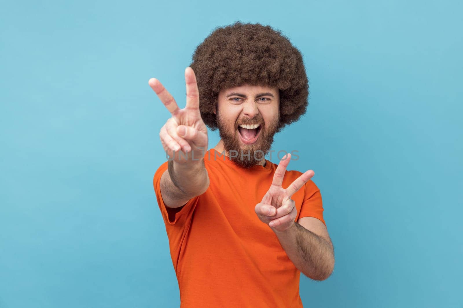 Portrait of excited cheerful handsome man with Afro hairstyle wearing orange T-shirt showing peace sign, v gesture and screaming happily. Indoor studio shot isolated on blue background.