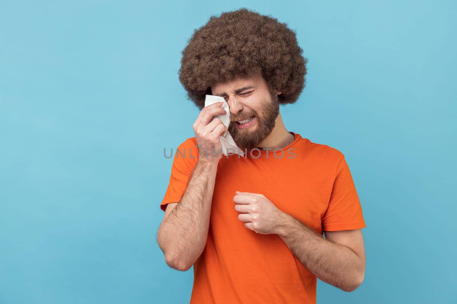 Portrait of depressed man with Afro hairstyle wearing orange T-shirt holding head down, hiding his face in hand and crying, feeling desperate. Indoor studio shot isolated on blue background.