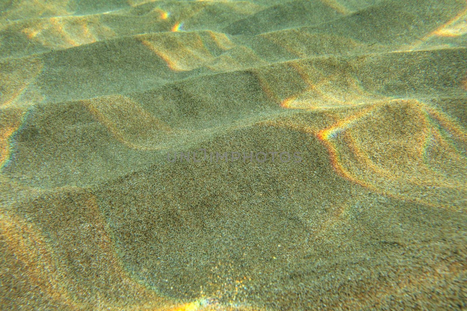 Underwater photo - sun shining on sand "dunes" in shallow water, casting small "rainbows". Abstract marine background.