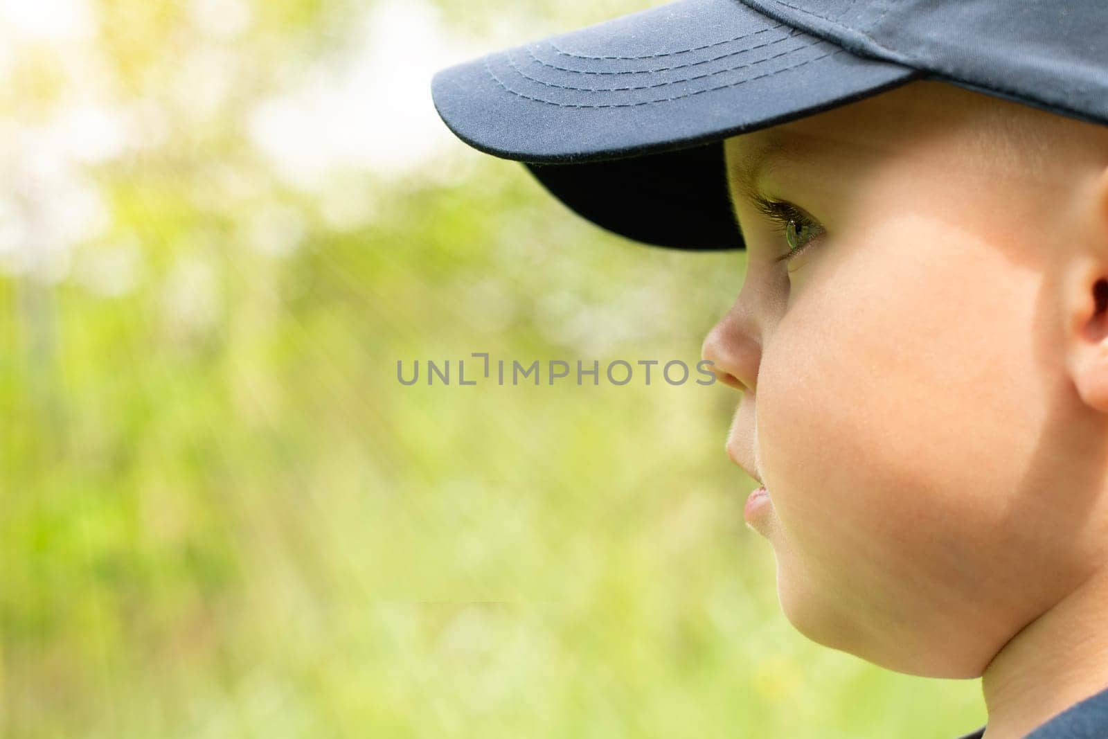 Face in profile of a little handsome boy in a blue cap on a green background in the sun. Close-up. copy space. Banner.
