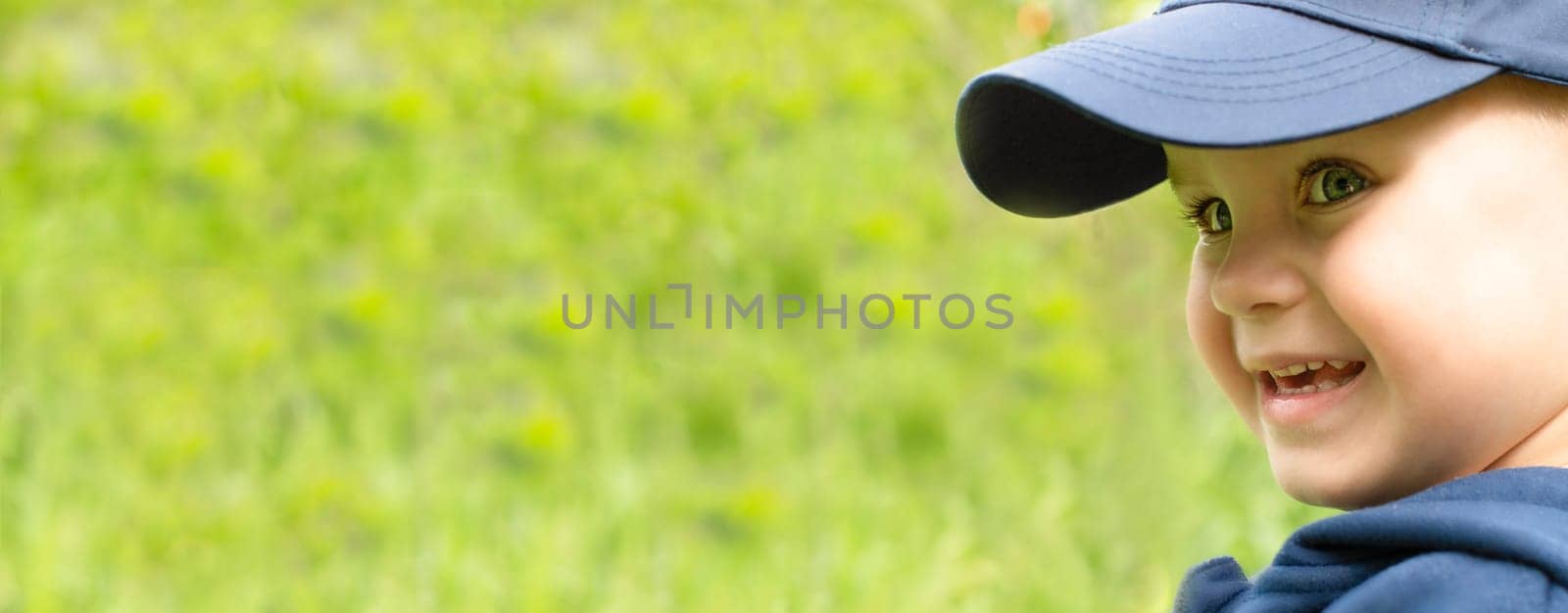 Face in profile of a little cheerful and handsome boy in a blue cap, smiling happily on a green background. close-up. Banner. copy space.