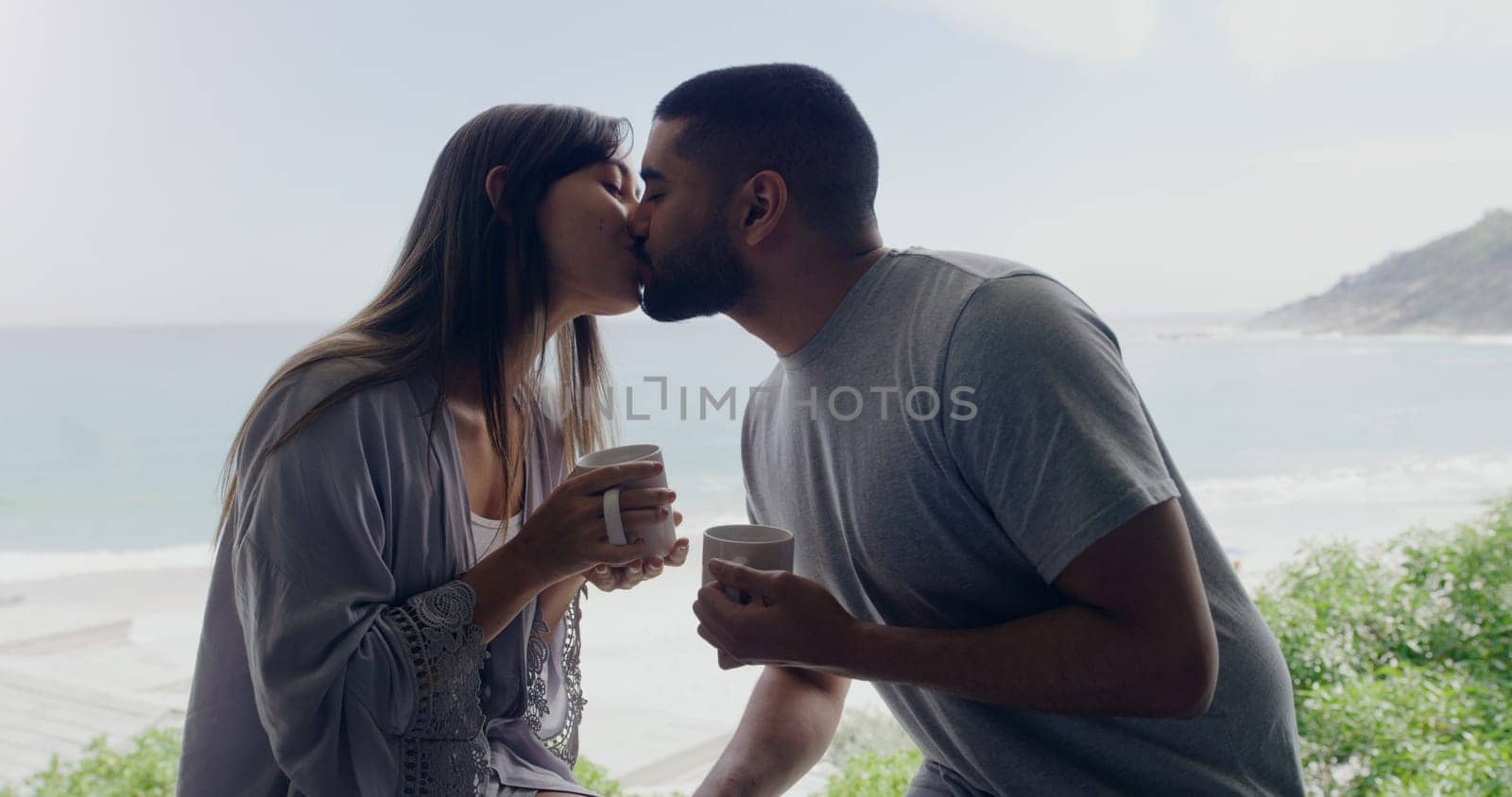 Couple, kiss and coffee in morning on balcony with love, romance and care on holiday by ocean with sunshine. Man, woman and trust with drink, espresso or matcha on vacation by sea with bond in summer by YuriArcurs