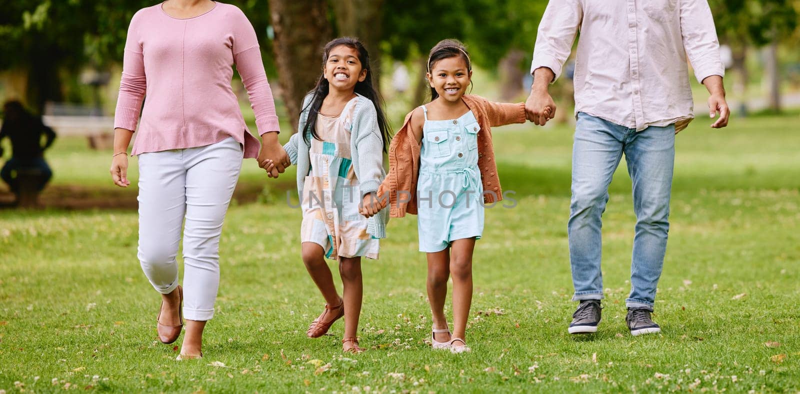 Family, park and children holding hands with parents walking, bonding and time in nature together with smile. Mother, father and kids walk on grass field for fun outdoor adventure in garden or woods.