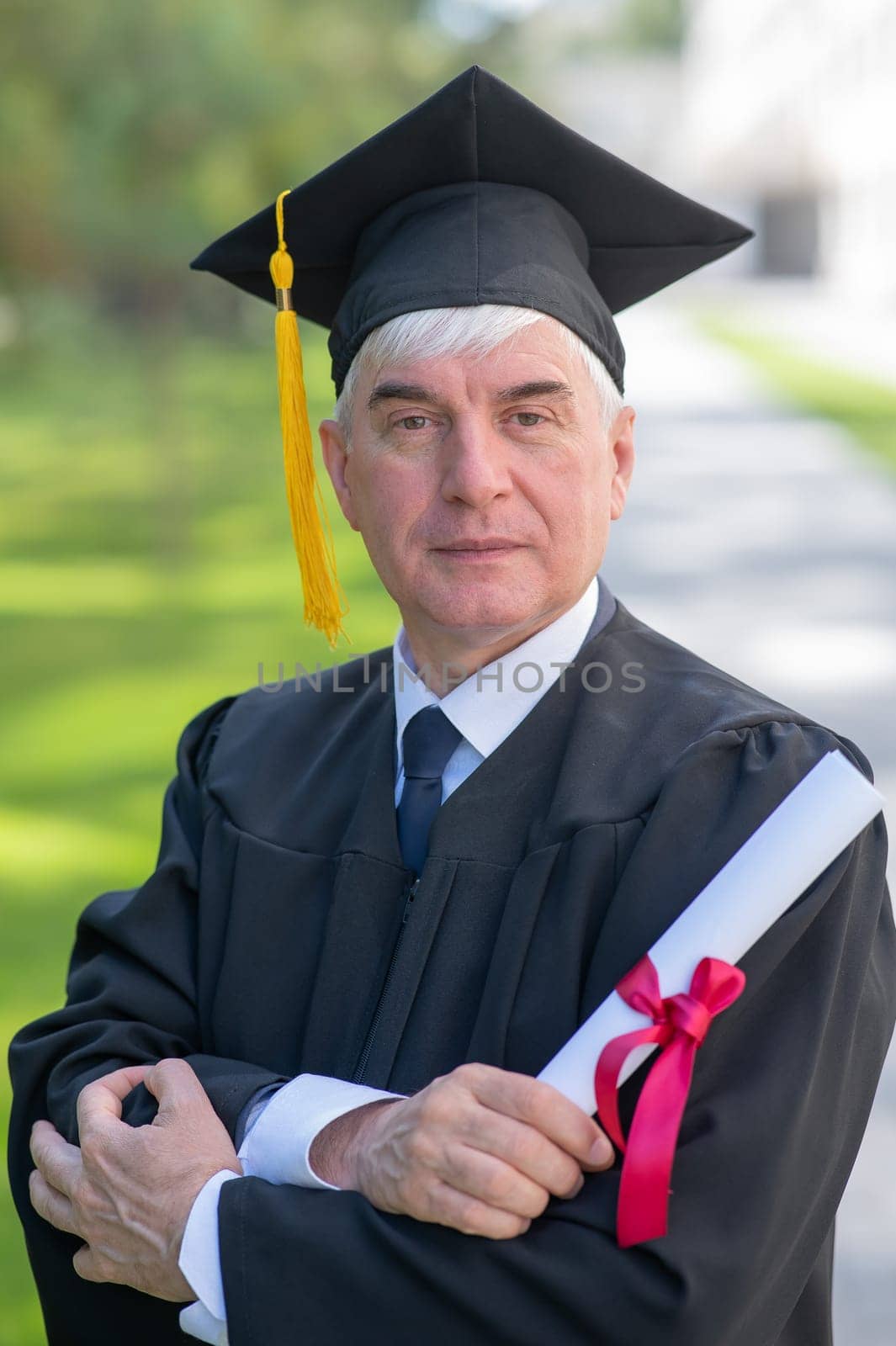 Portrait of an elderly man in a graduation gown and with a diploma in his hands outdoors. Vertical