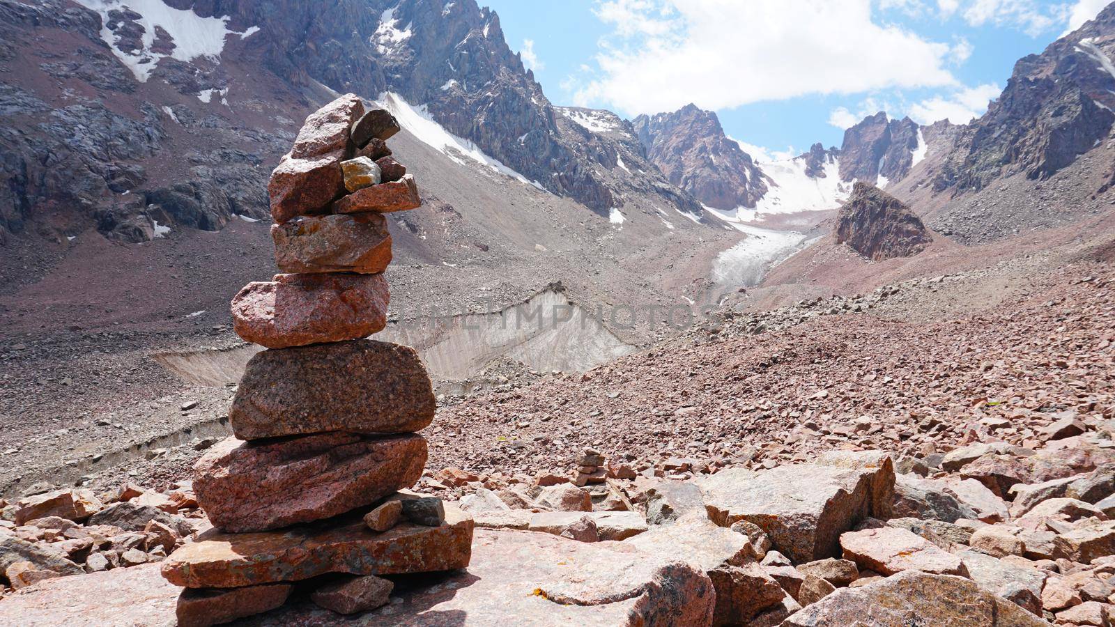 A pyramid of stones as a pointer in the mountains. Big rocks, high peaks and a snowy glacier. A moraine lake can be seen in the distance. The ice is covered with huge rocks. A cairn.