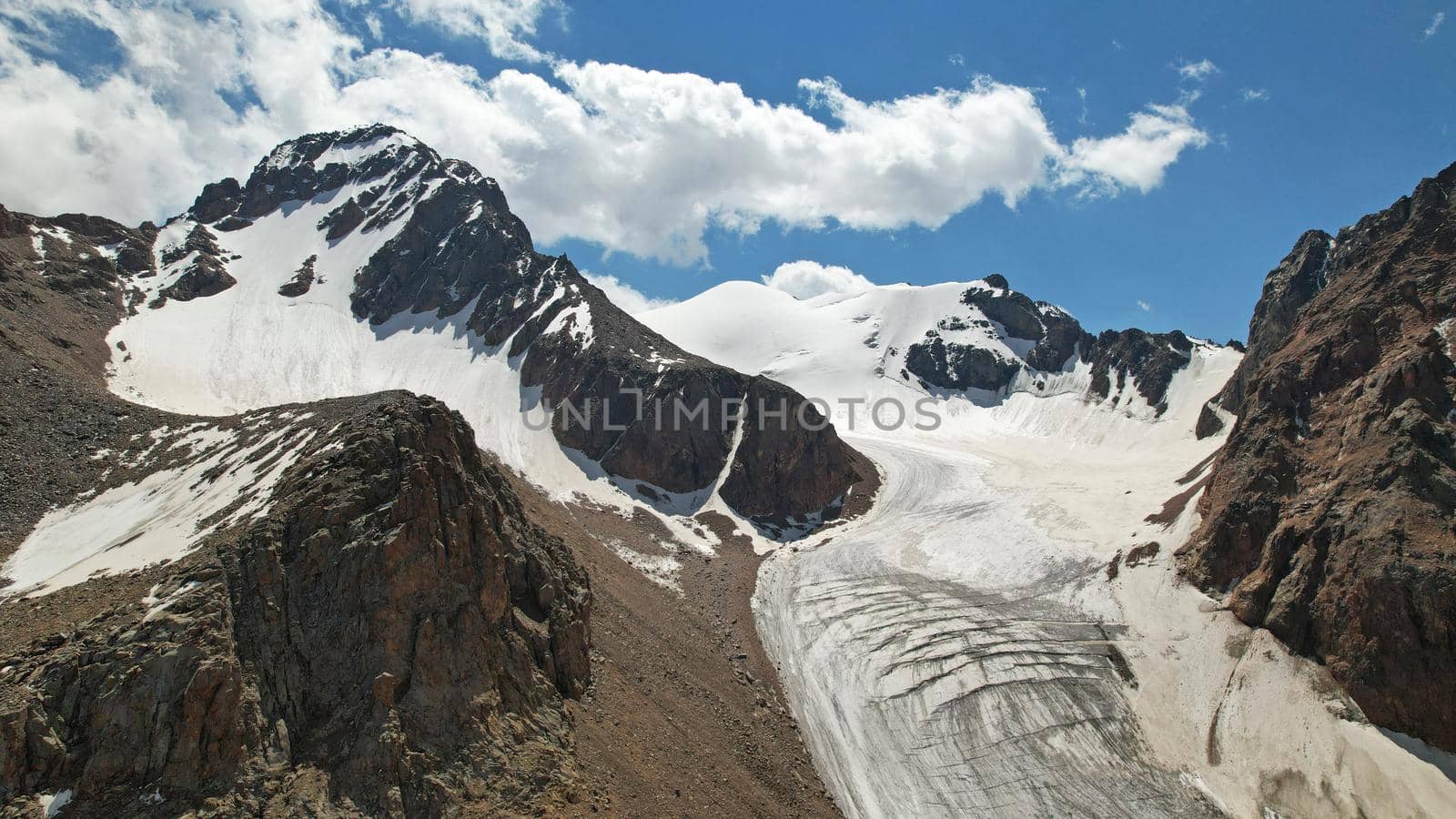 High rocky mountains are sometimes covered with ice and snow. A huge glacier passes between the peaks. The ice is gradually melting. The stones are lying on glacier. Steep cliffs. The view from drone