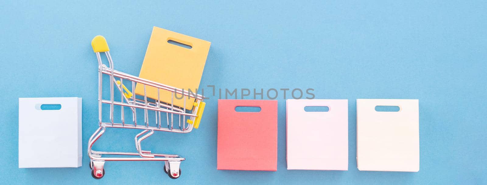 Abstract design element, concept of annual sale, shopping season - mini yellow cart with paper bag isolated on pastel blue background, top view, flat lay. by ROMIXIMAGE