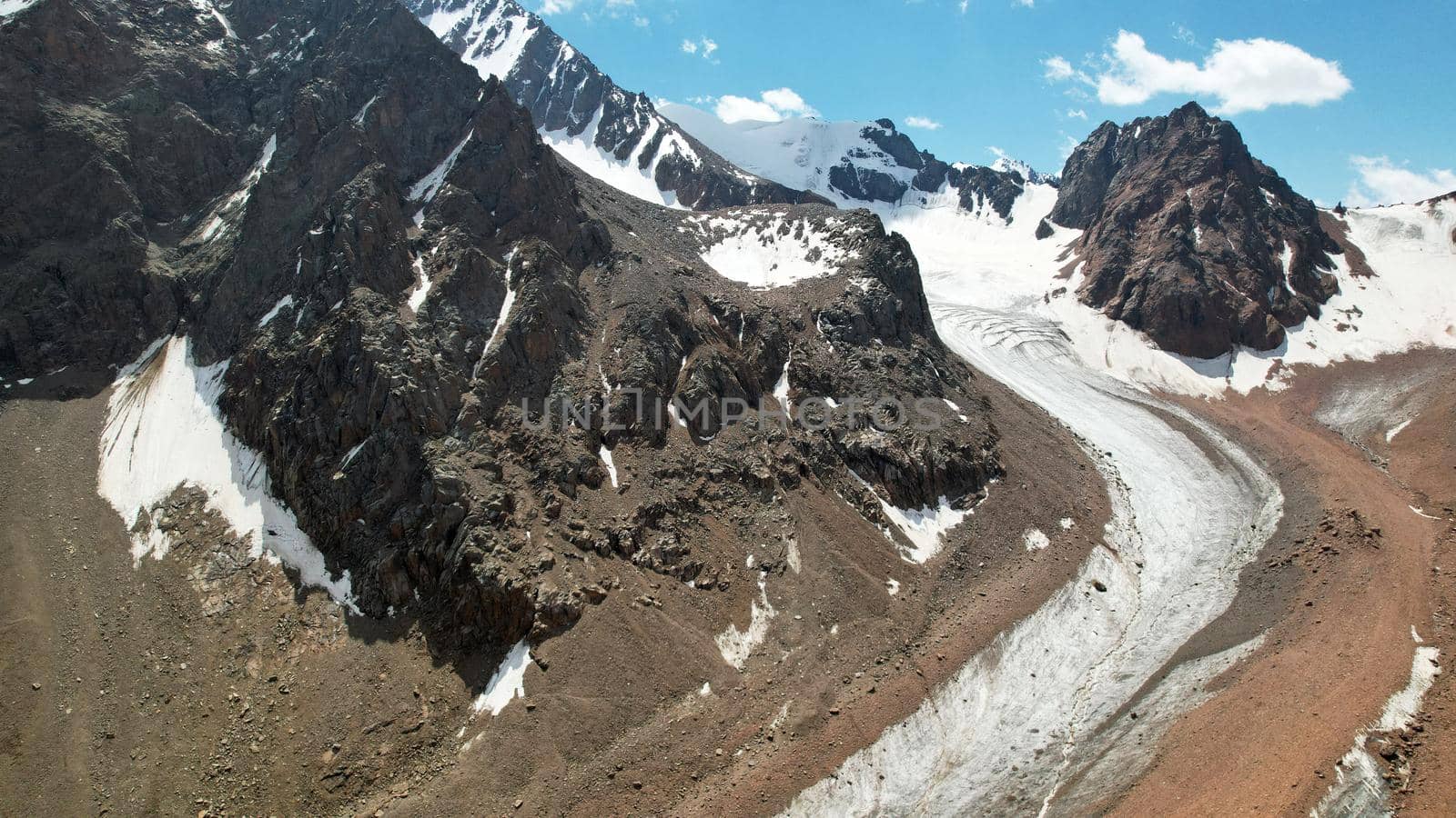 High rocky mountains are sometimes covered with ice and snow. A huge glacier passes between the peaks. The ice is gradually melting. The stones are lying on glacier. Steep cliffs. The view from drone