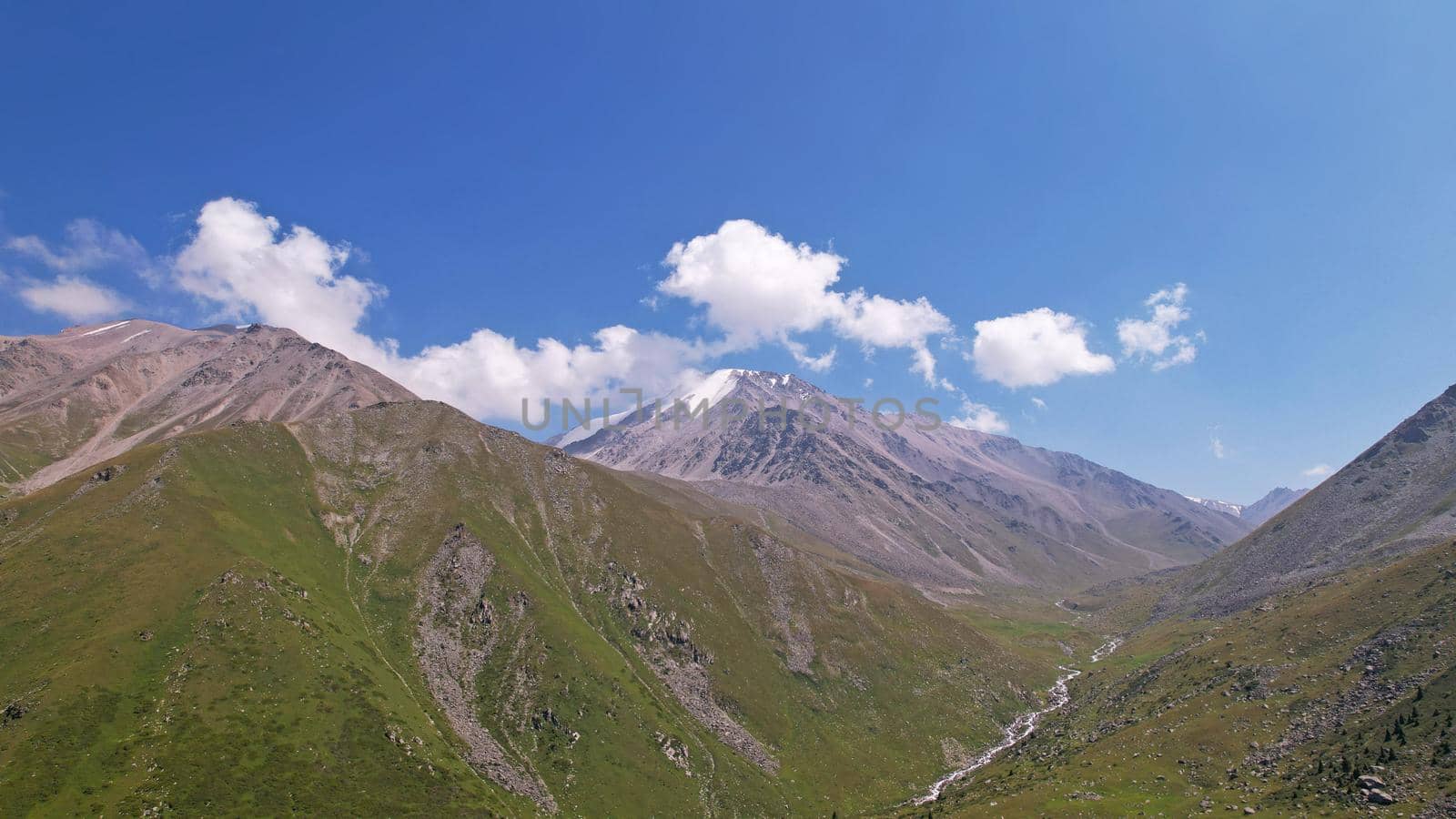 Clouds flow around the tops of high mountains. Rocky gorges and cliffs. The sky is blue, the sun is shining brightly, green grass is growing in places. Mountain terrain from the height of the drone