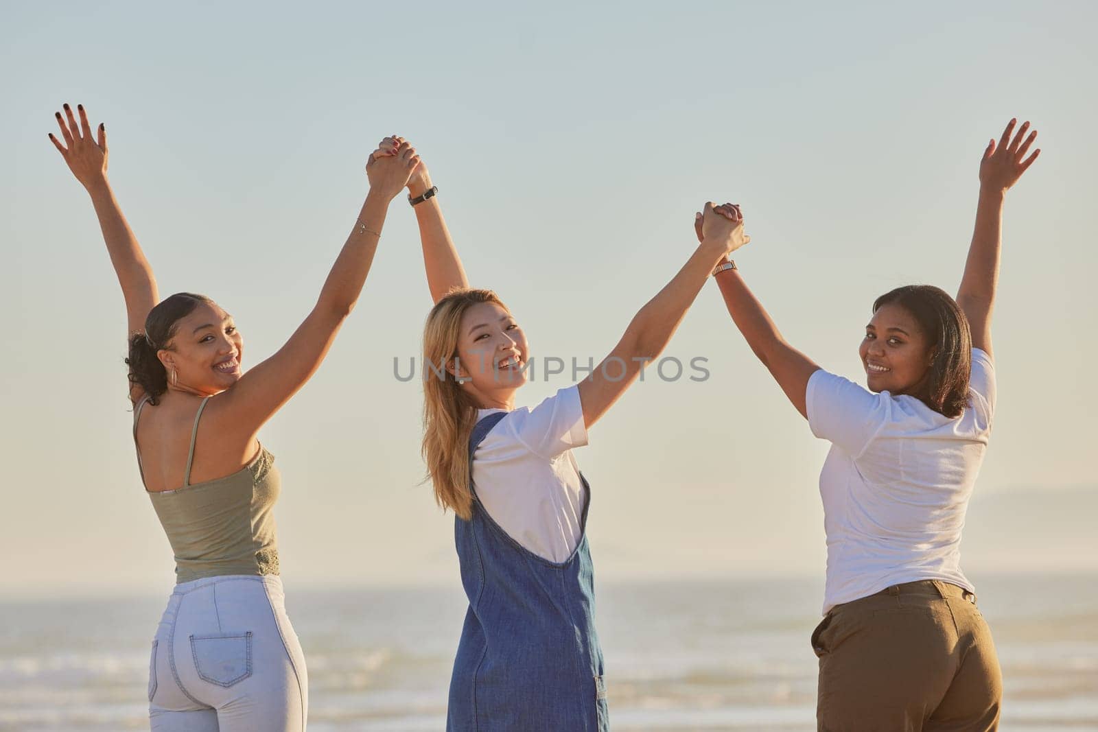 Happy, girl friends and holding hands at a beach with women on a holiday, freedom and travel. Portrait of people with arms raised to show excited adventure by the ocean and sea together by water.