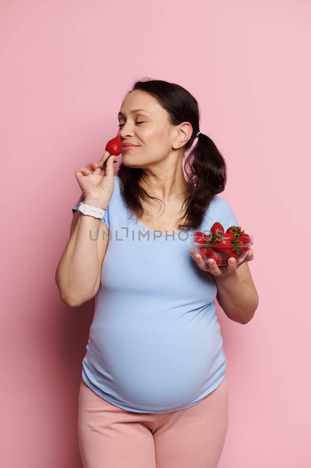 Beautiful glamorous pregnant woman in a blue t-shirt and pink pants, sniffing fresh ripe strawberries, enjoying a healthy diet during pregnancy, posing with bowl of berries on isolated pink background