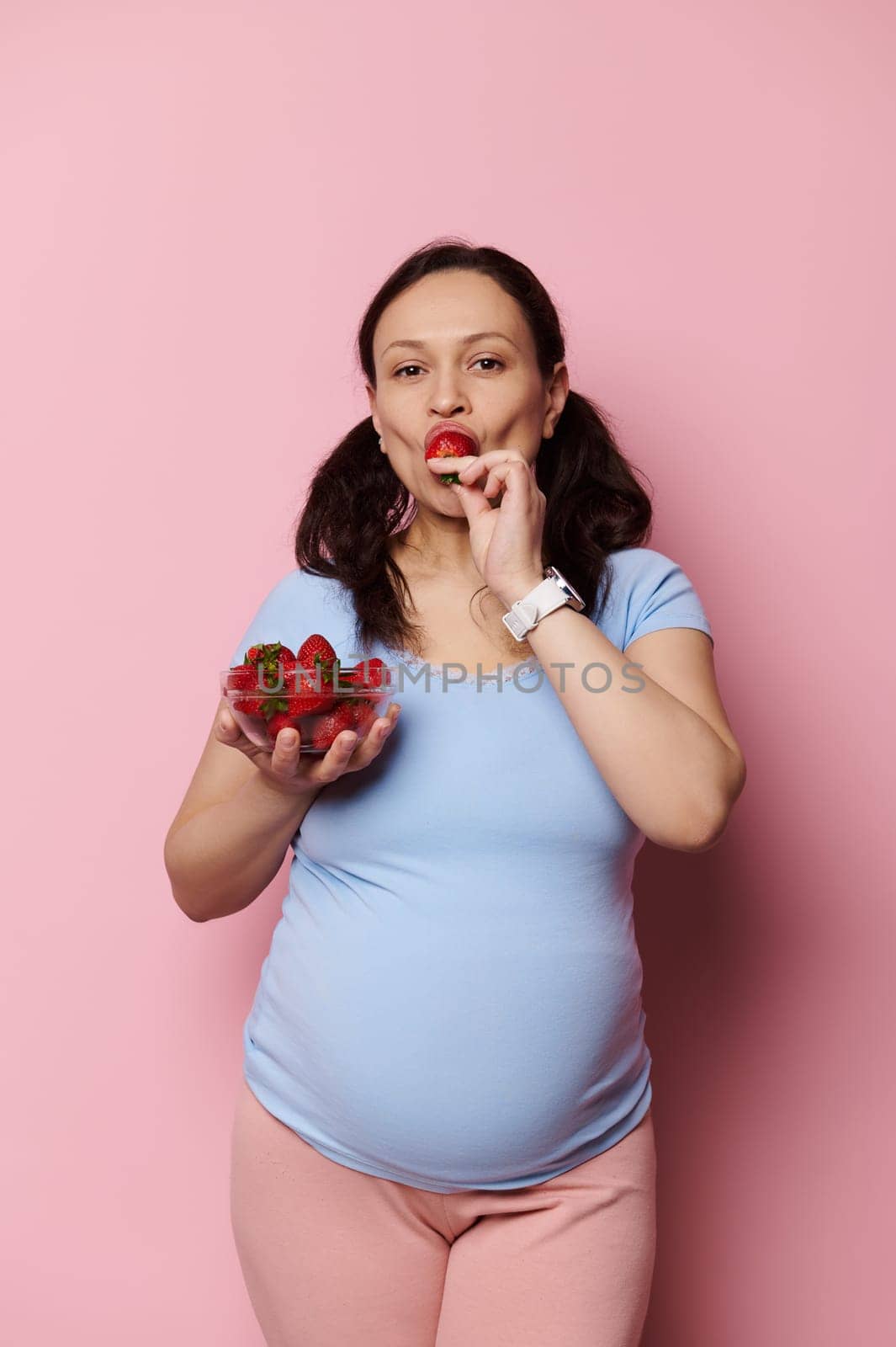 Vertical advertising studio shot on pink background of an expectant pregnant mother in blue t-shirt, looking at camera, holding a bowl with fresh berries and tasting yummy strawberries. Healthy diet