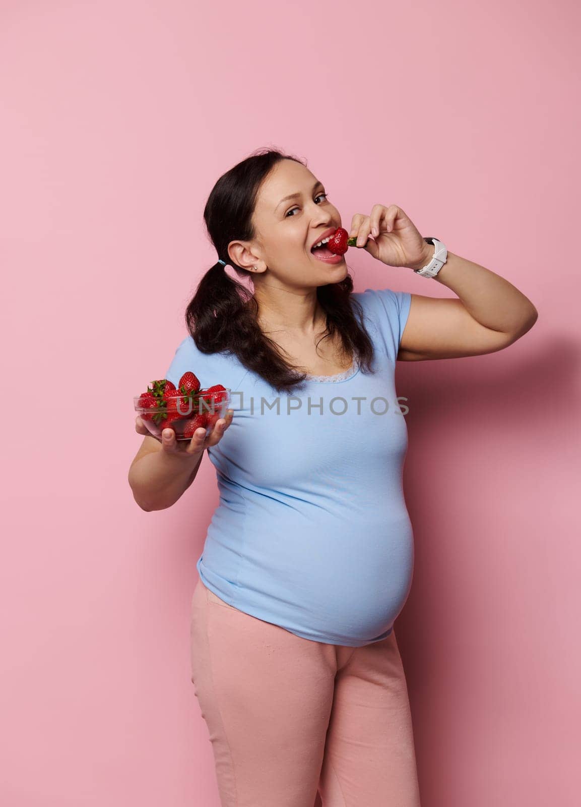 Gorgeous pregnant woman eating fresh organic strawberries, isolated over pink background. Expectant mother in blue t-shirt, holding bowl with fresh berries. Healthy nutrition and pregnancy concept.