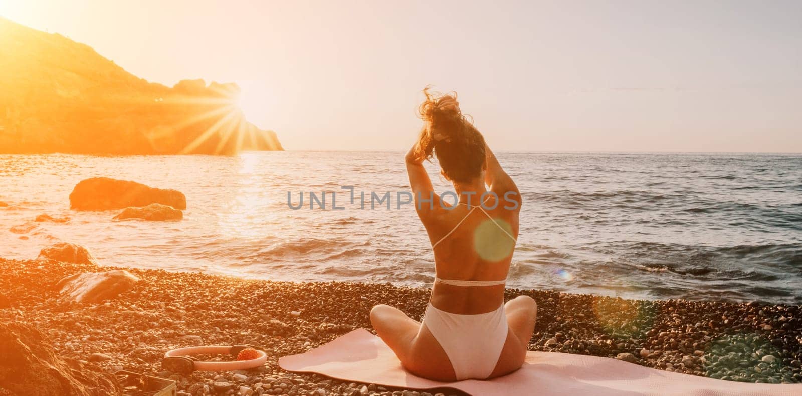 Young woman in swimsuit with long hair practicing stretching outdoors on yoga mat by the sea on a sunny day. Women's yoga fitness pilates routine. Healthy lifestyle, harmony and meditation concept.