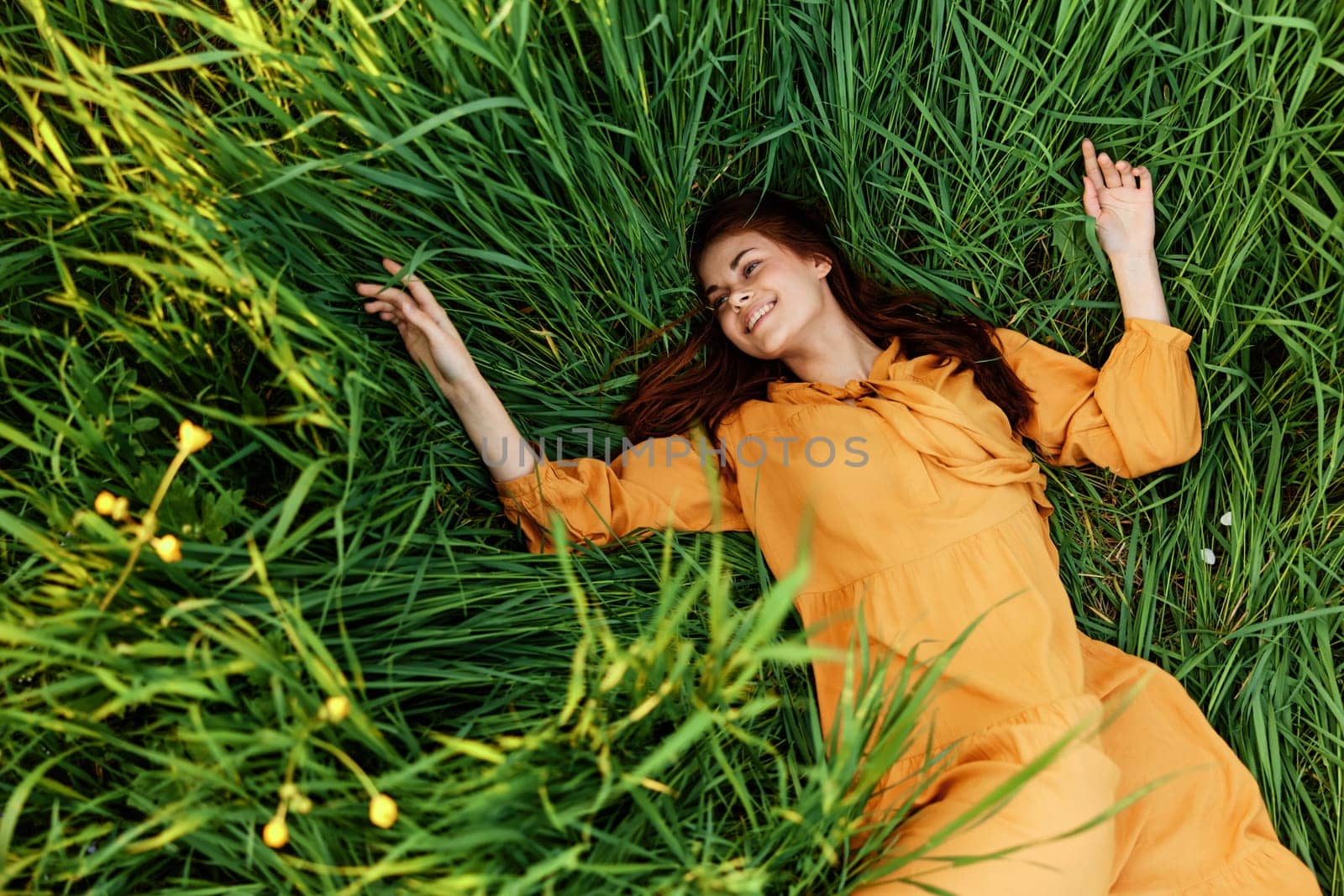 a relaxed red-haired woman enjoys summer lying in the tall green grass in a long orange dress smiling happily with her eyes closed. High quality photo