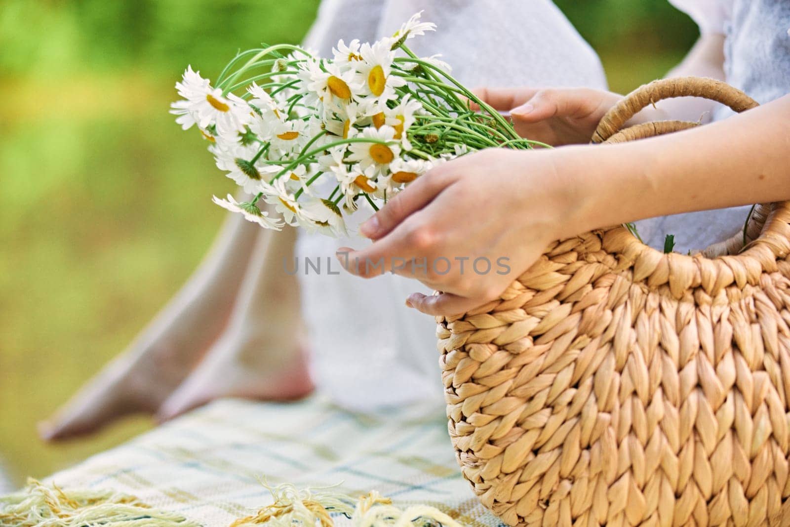 a woman holds a wicker bag with daisies with her hand while sitting on the pier by Vichizh