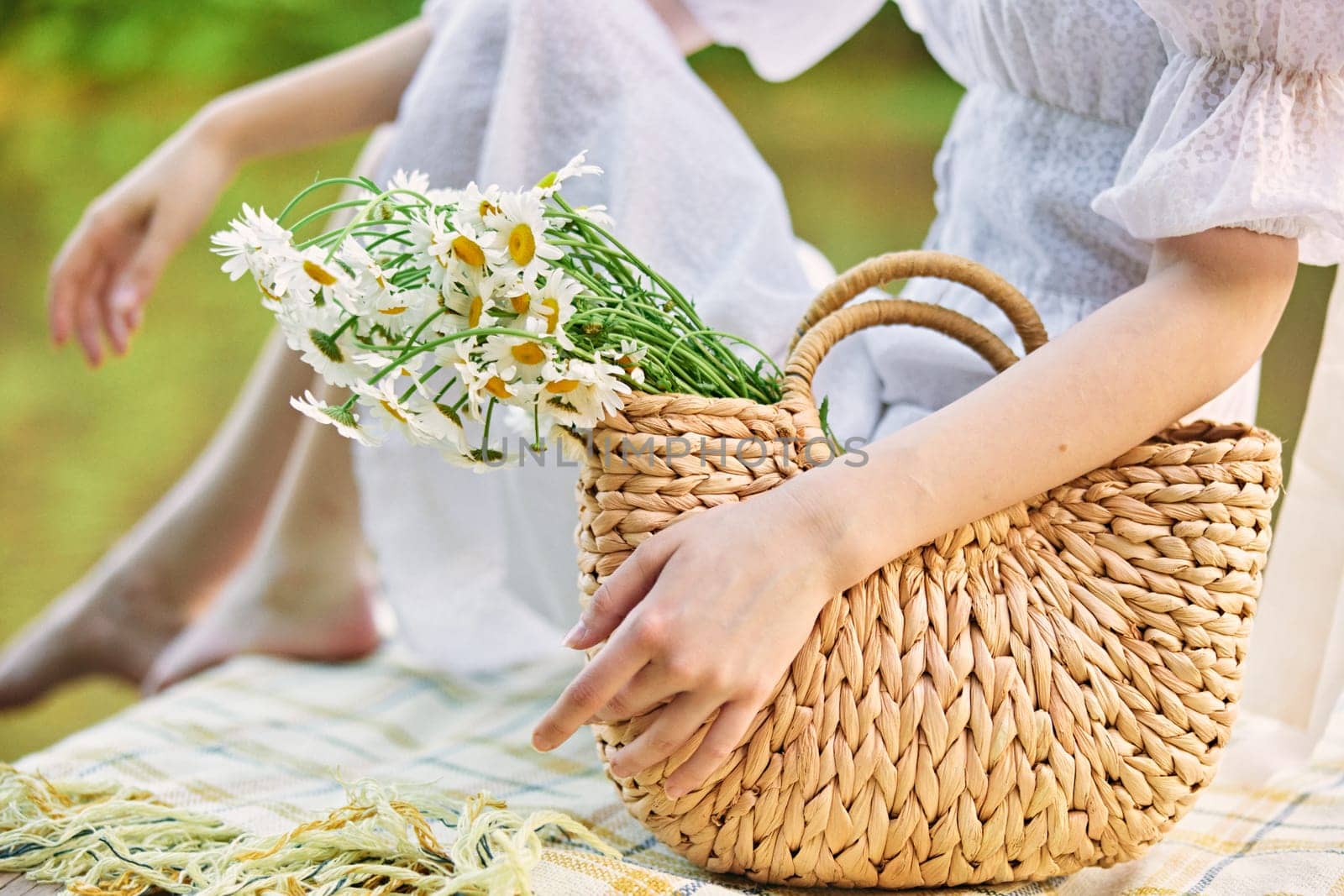 a woman holds a wicker bag with daisies with her hand while sitting on the pier. High quality photo