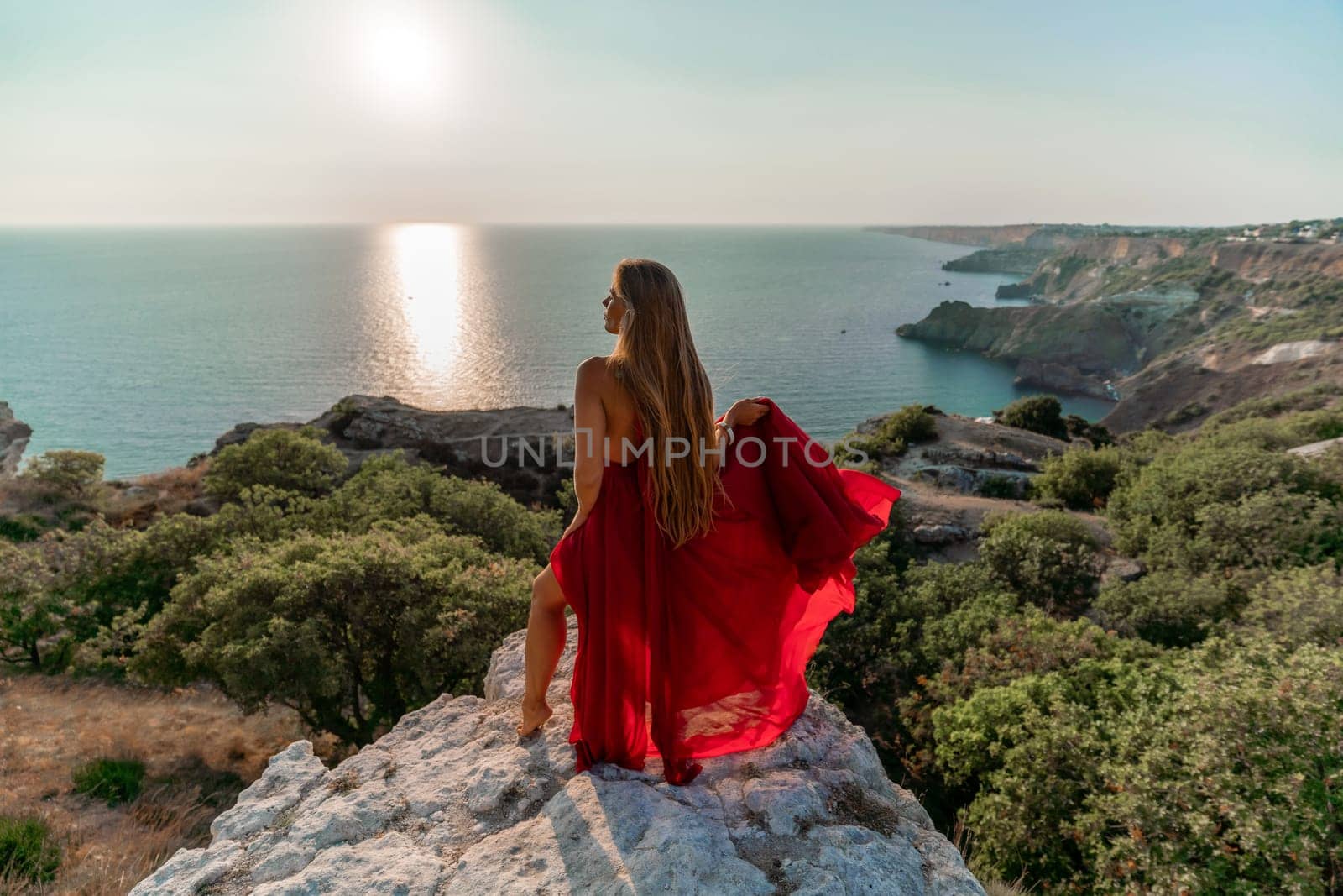 Woman sunset sea red dress, back view a happy beautiful sensual woman in a red long dress posing on a rock high above the sea on sunset