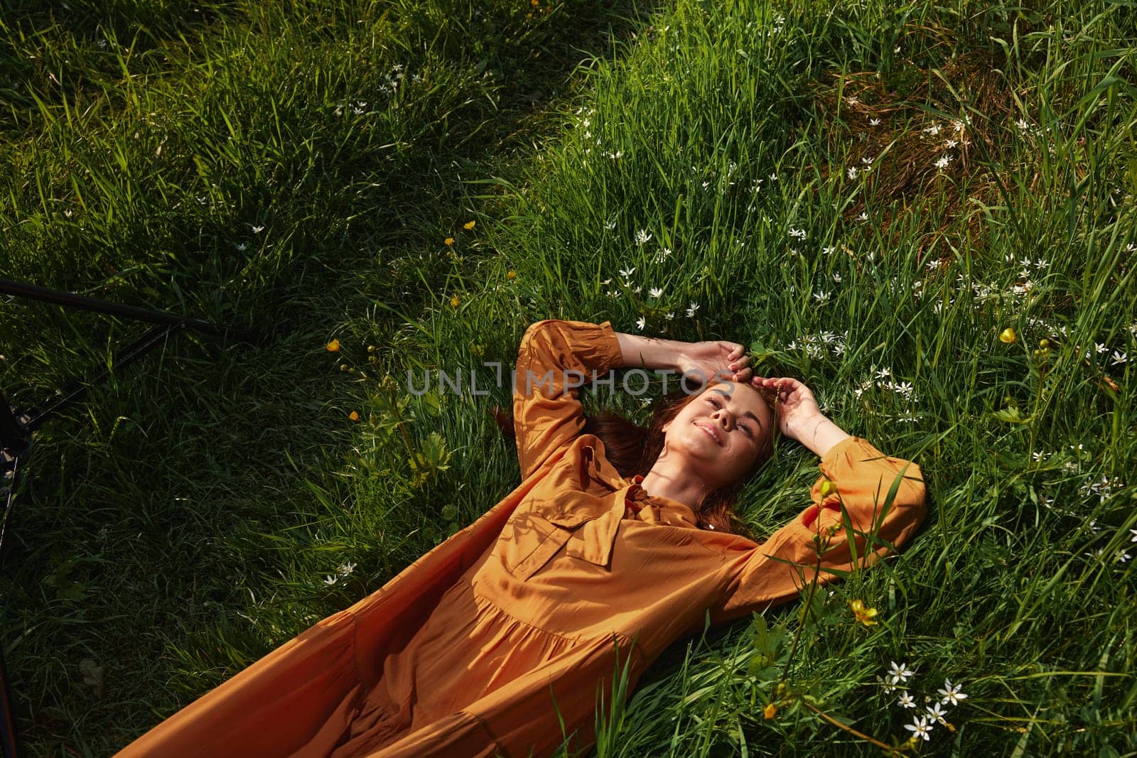 a happy, relaxed woman, resting lying in the green grass, in a long orange dress, with her eyes closed and a pleasant smile on her face, enjoying harmony with nature and recuperating by Vichizh