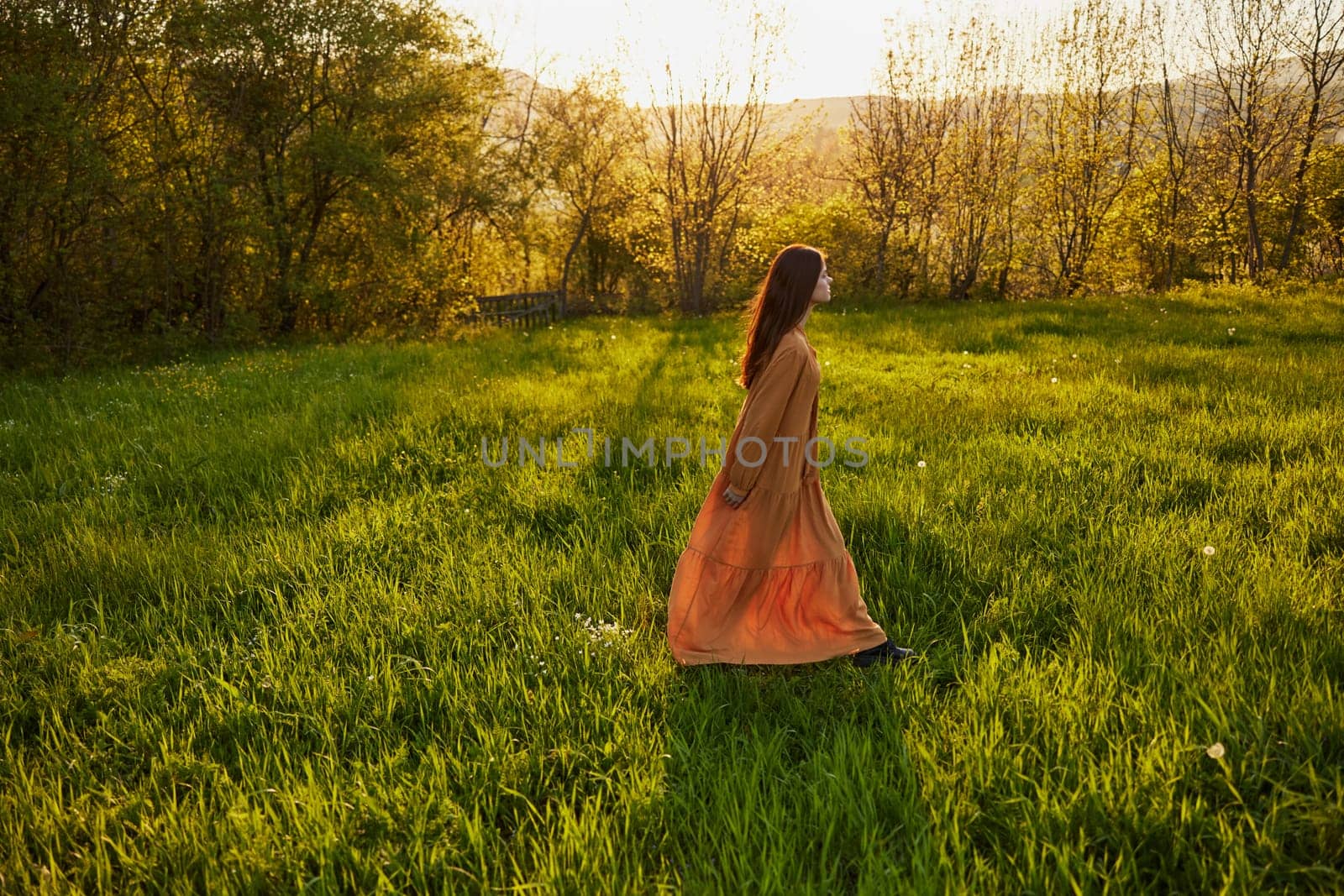 an attractive, slender, red-haired woman stands in a wide green field during sunset in a long orange dress enjoying unity with nature and relaxation. High quality photo