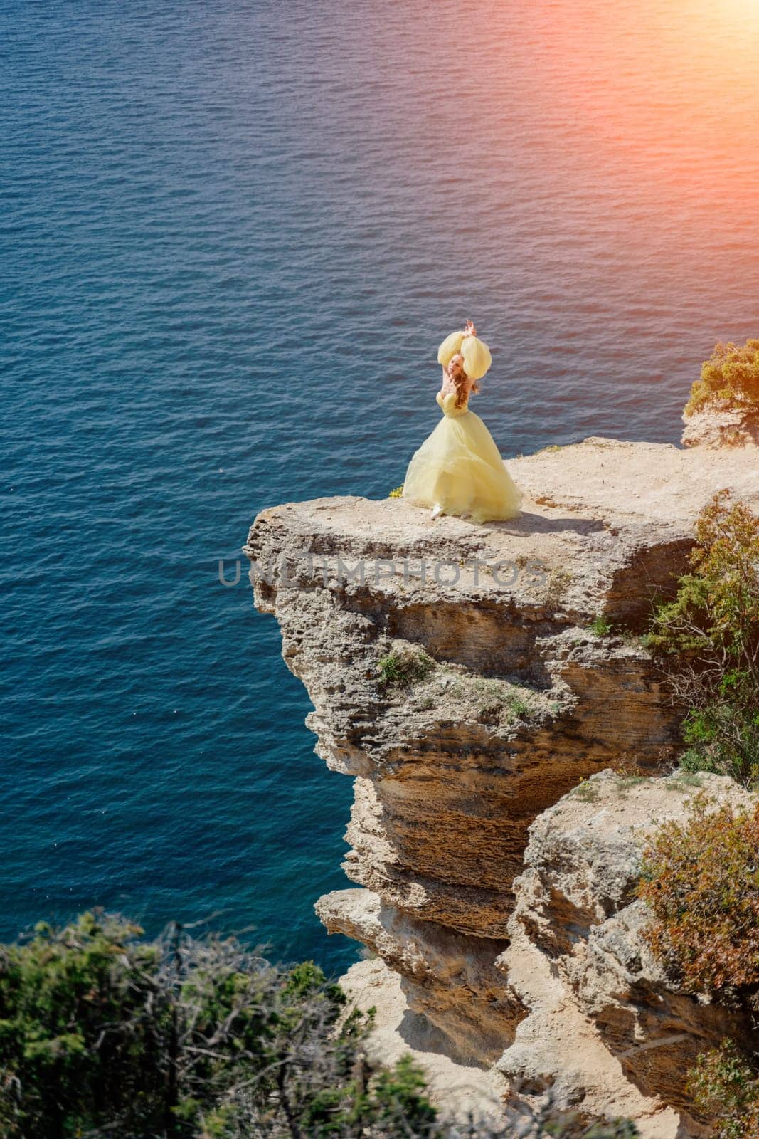 Woman yellow dress sea. Side view Young beautiful sensual woman in yellow long dress posing on a rock high above the sea at sunset. Girl in nature against the blue sky by Matiunina