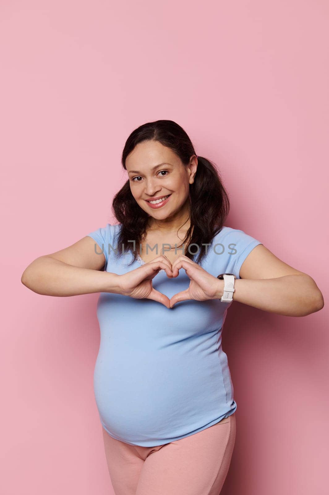 Joyful pregnant woman in blue t-shirt, gestures with hands, showing heart shape with fingers, smiles broadly looking at camera, isolated pink background. Pregnancy trimester third. Maternity lifestyle
