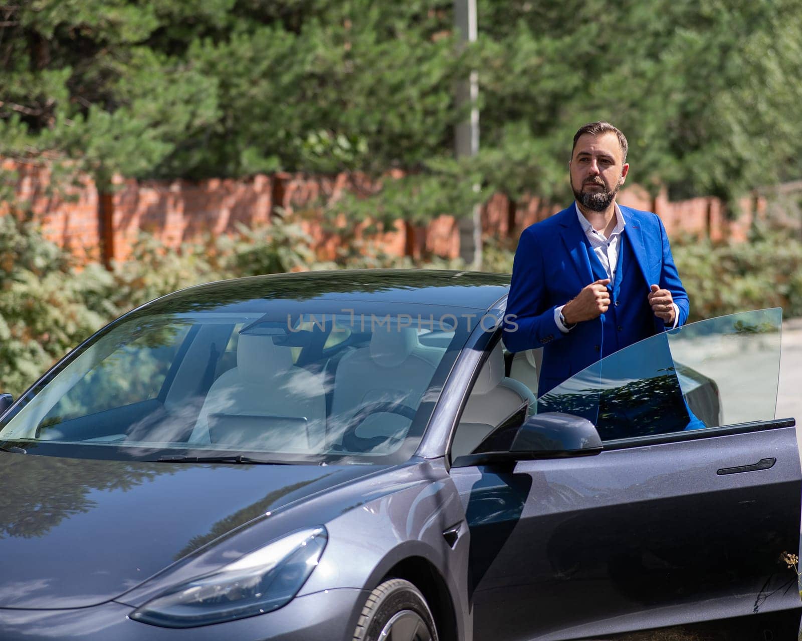 Caucasian bearded man in a blue suit gets out of a black electro car in the countryside in summer