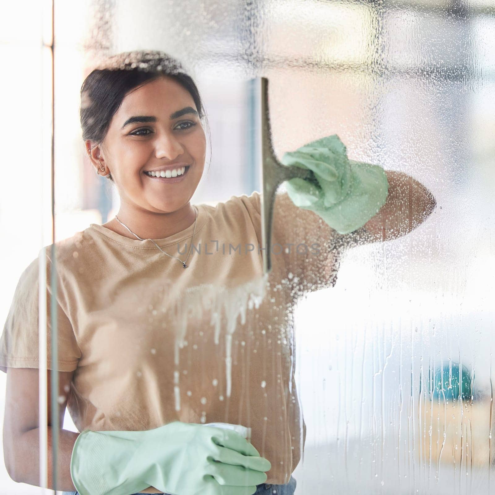 Happy, smile and girl cleaning window with spray bottle and soap or detergent, housekeeper in home or hotel. Housework, smudge and woman or professional cleaner service washing off glass in apartment.