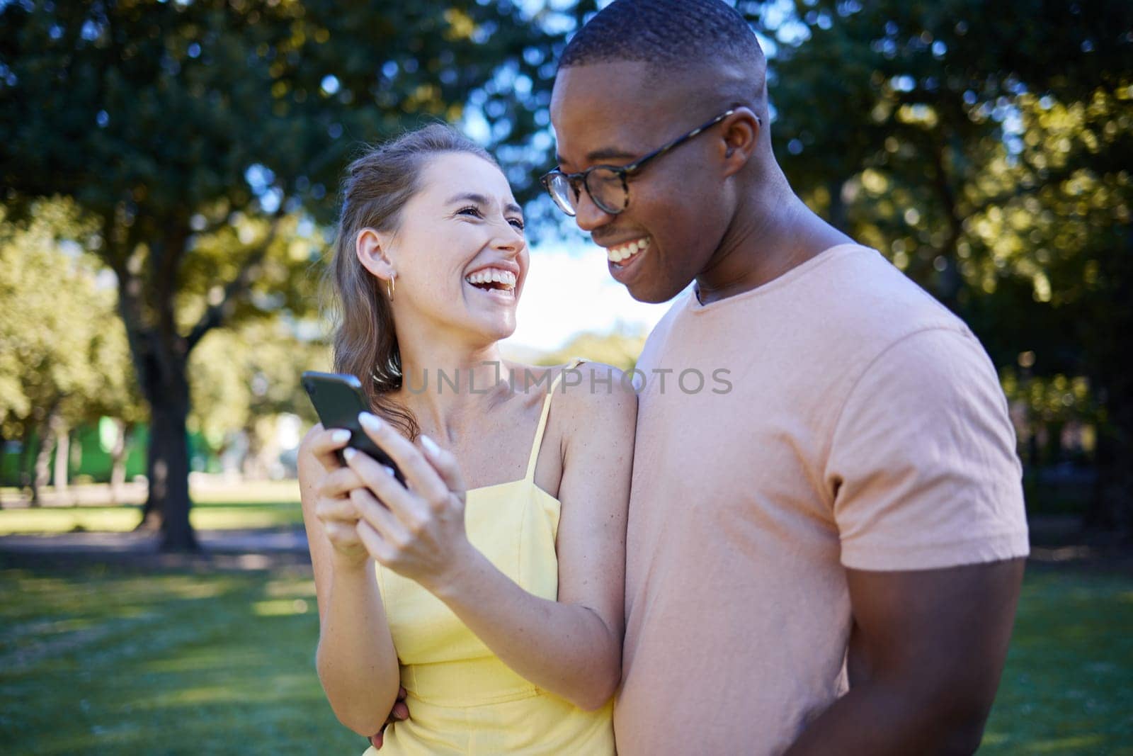 Interracial, social media and couple with a phone in nature, funny communication and laughing at meme. Comic, streaming and black man and woman reading a joke on a mobile in a park in France by YuriArcurs