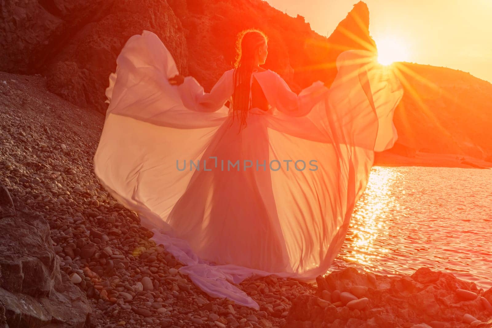 Mysterious woman silhouette long hair walks on the beach ocean water, sea nymph wind listens to the wave. Throws up a long white dress, a divine sunset. Artistic photo from the back without a face.