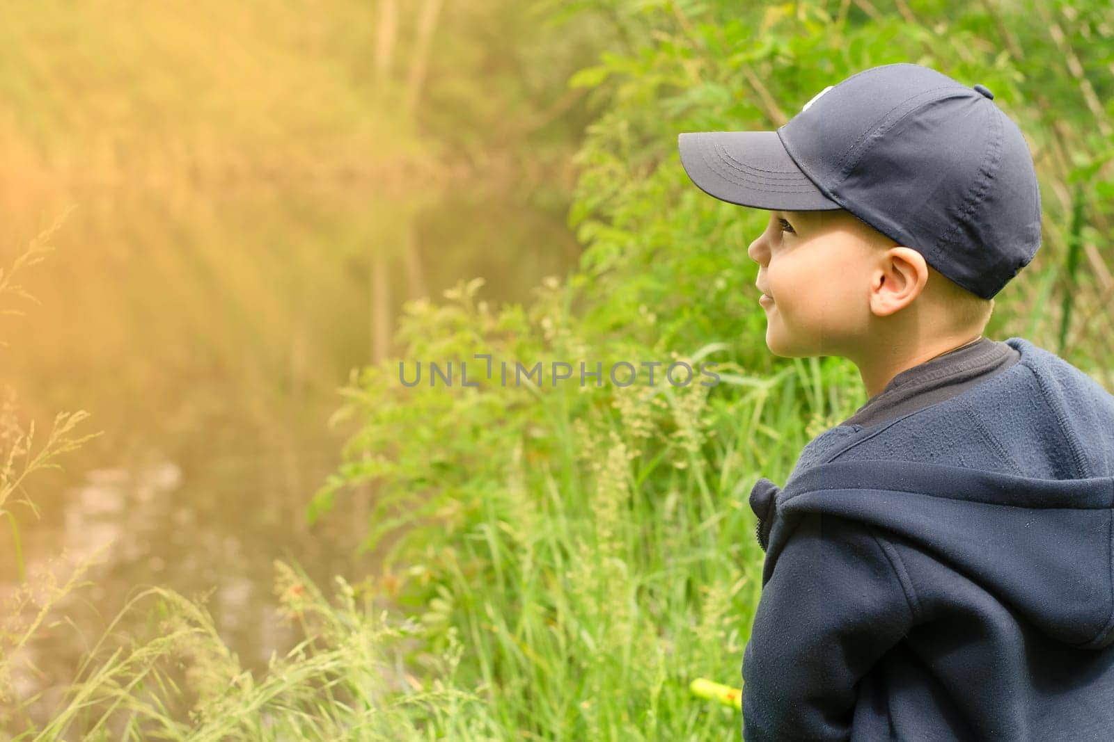 A little handsome boy in a blue cap stands in profile near a lake in a green forest in the rays of the setting sun and laughs happily. copy space.