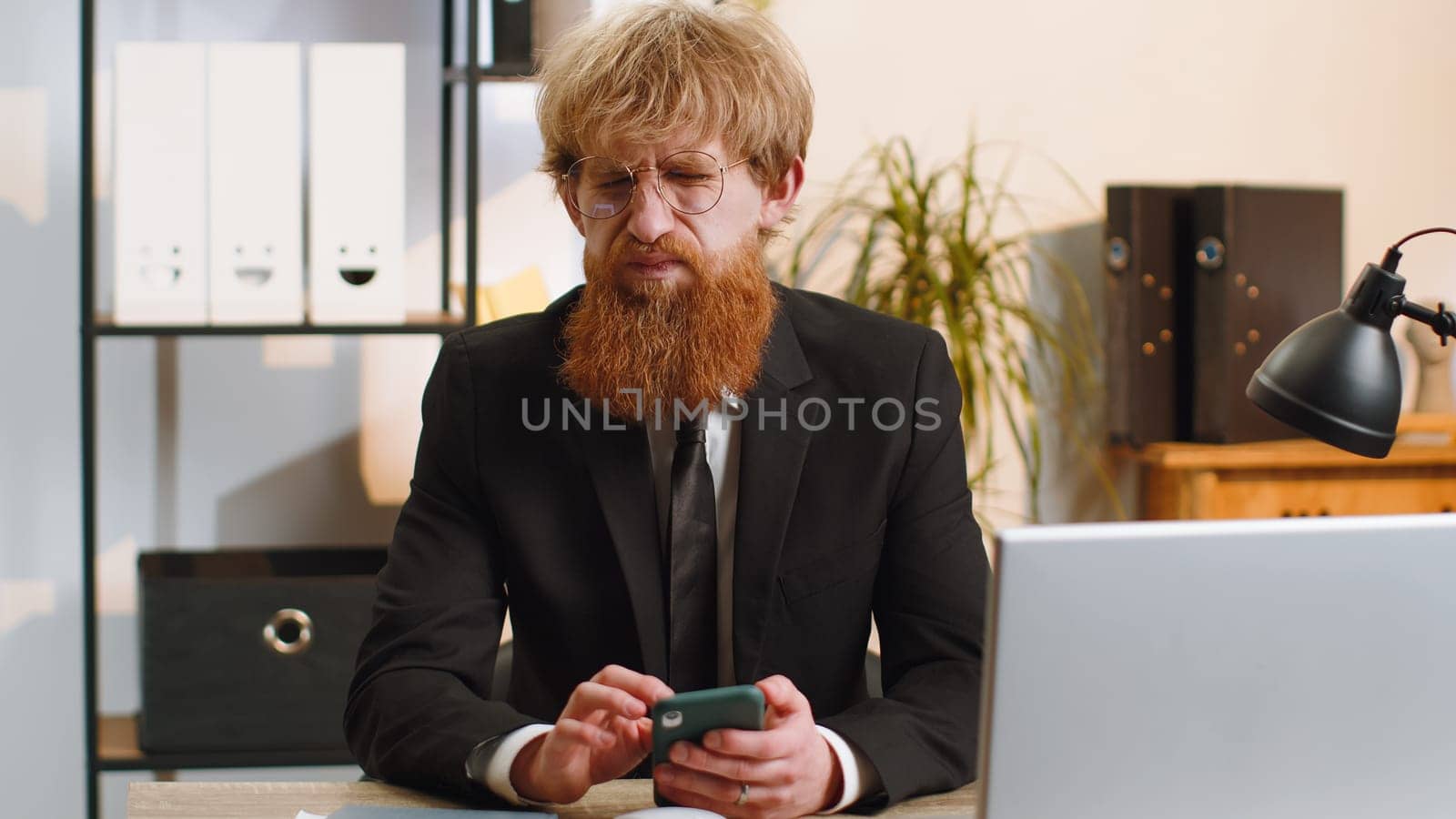 Young businessman in suit use smartphone typing browsing, loses becoming surprised sudden lottery results, bad news, fortune loss fail at home office desk workplace. Bearded freelancer worker man