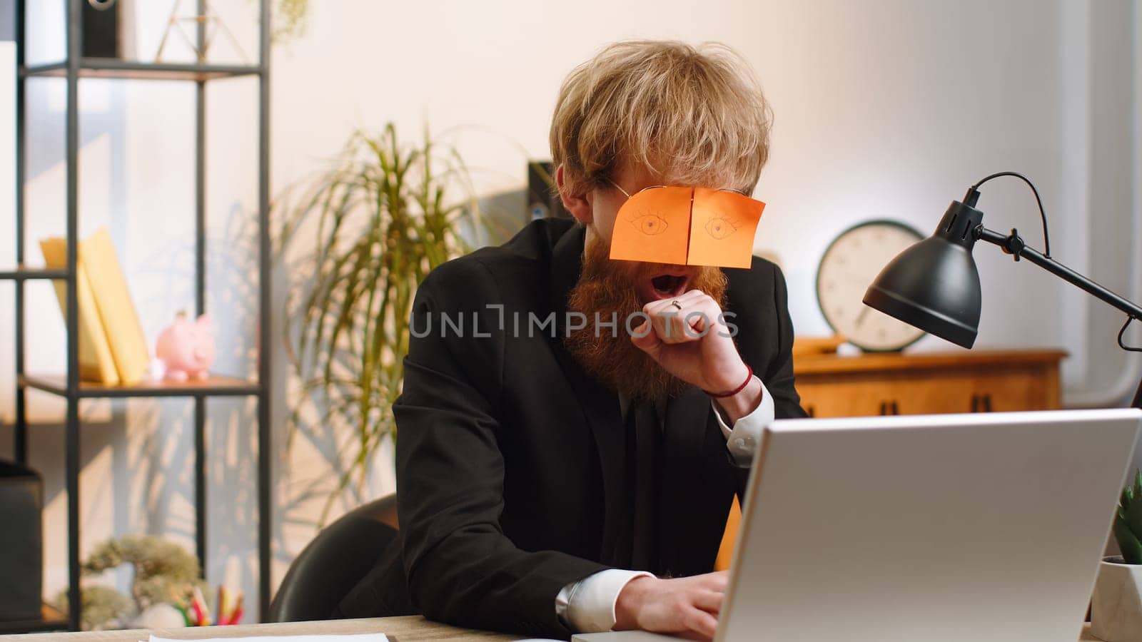 Inefficient tired young businessman working sleeping on laptop computer with eyes stickers on face at office workplace desk. Bearded lazy manager freelancer man. Business people cheating to sleep