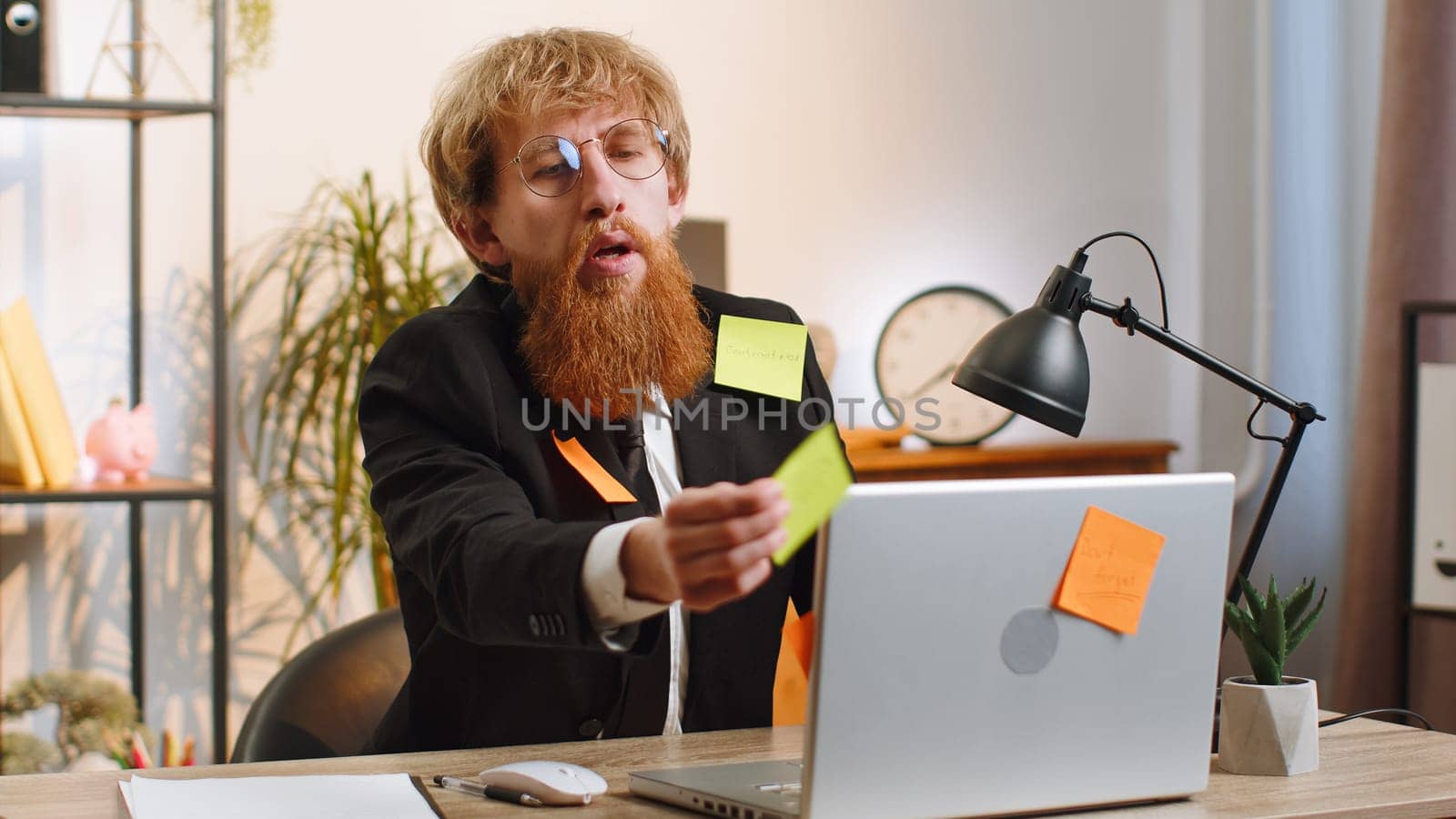 Tired exhausted young businessman working on laptop at office with many sticker task inscriptions. Freelancer man busy occupation feels sad boredom overworked multitasking. Panic attack, deadline