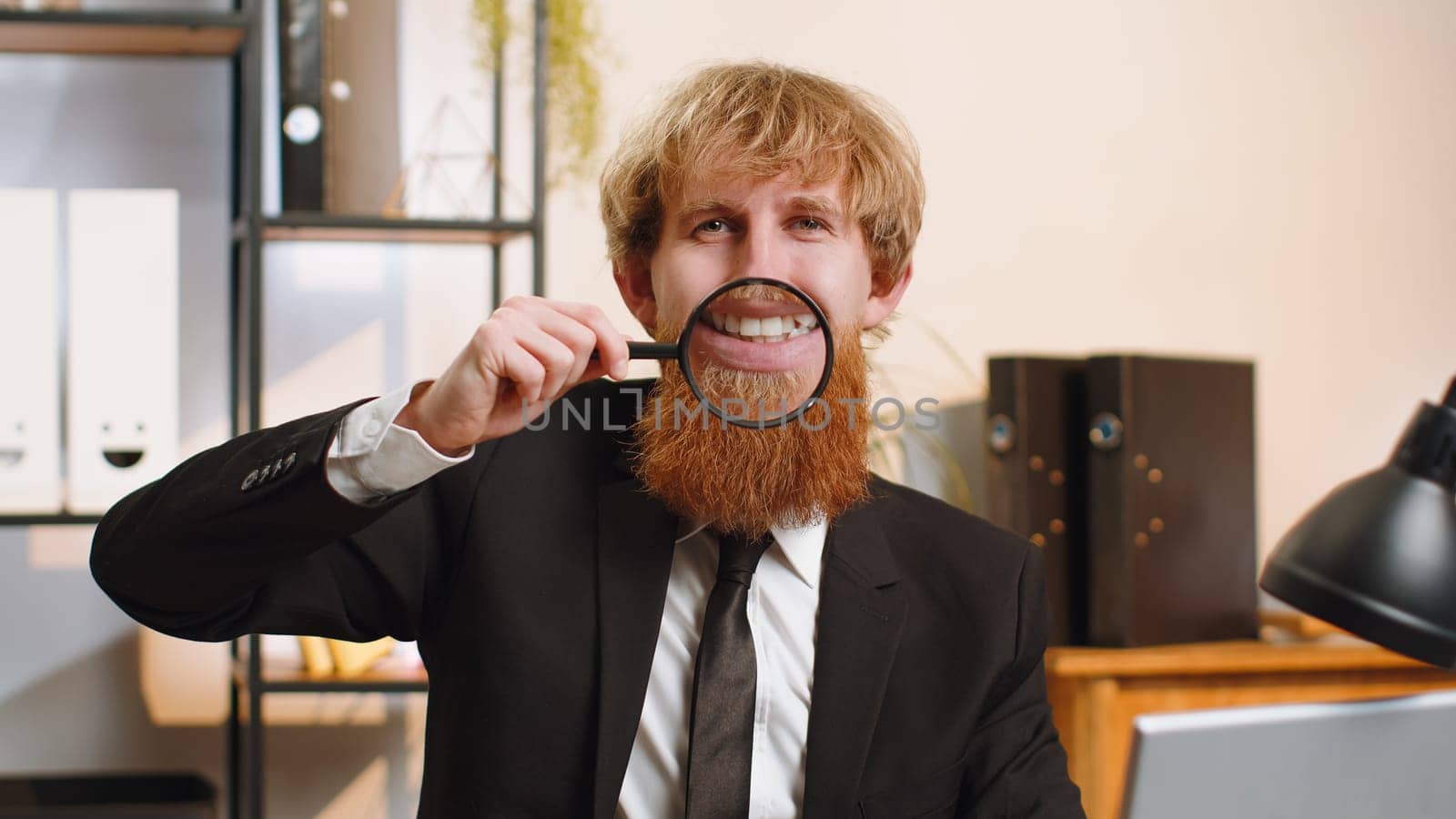 Young businessman guy working at home office desk workplace holding magnifier glass on teeth, looking at camera with happy expression, showing funny smiling silly face mouth. Manager freelancer man