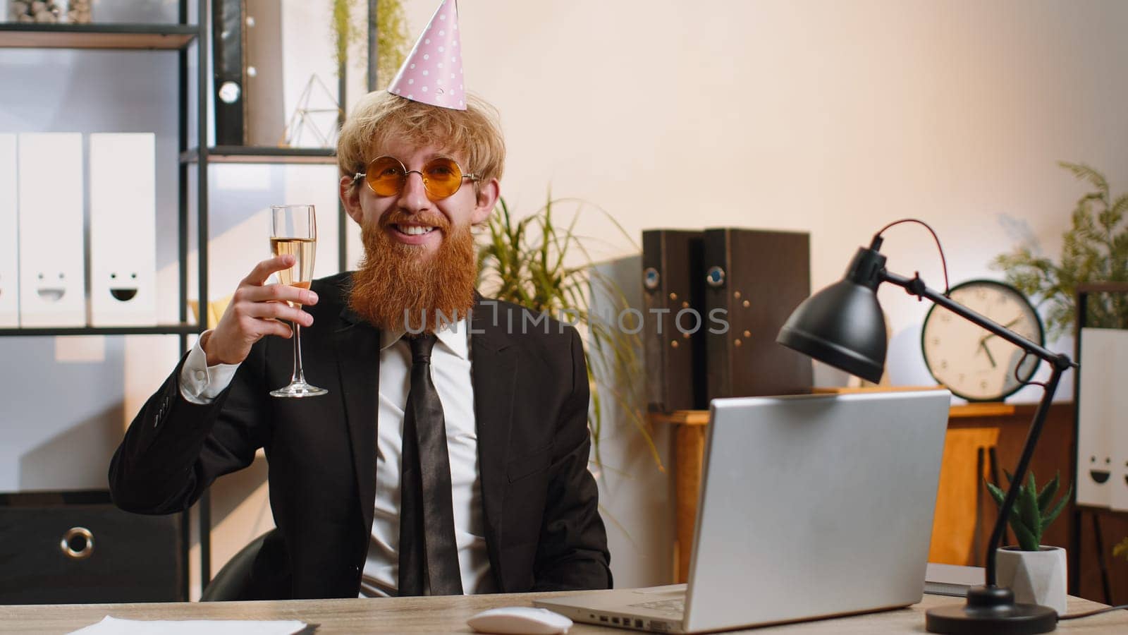 Young business man wears sunglasses, festive cap, drinking champagne at workplace dancing victory dance, weekend. Rejoicing developing project. Celebrating success. Lottery jackpot. Office people