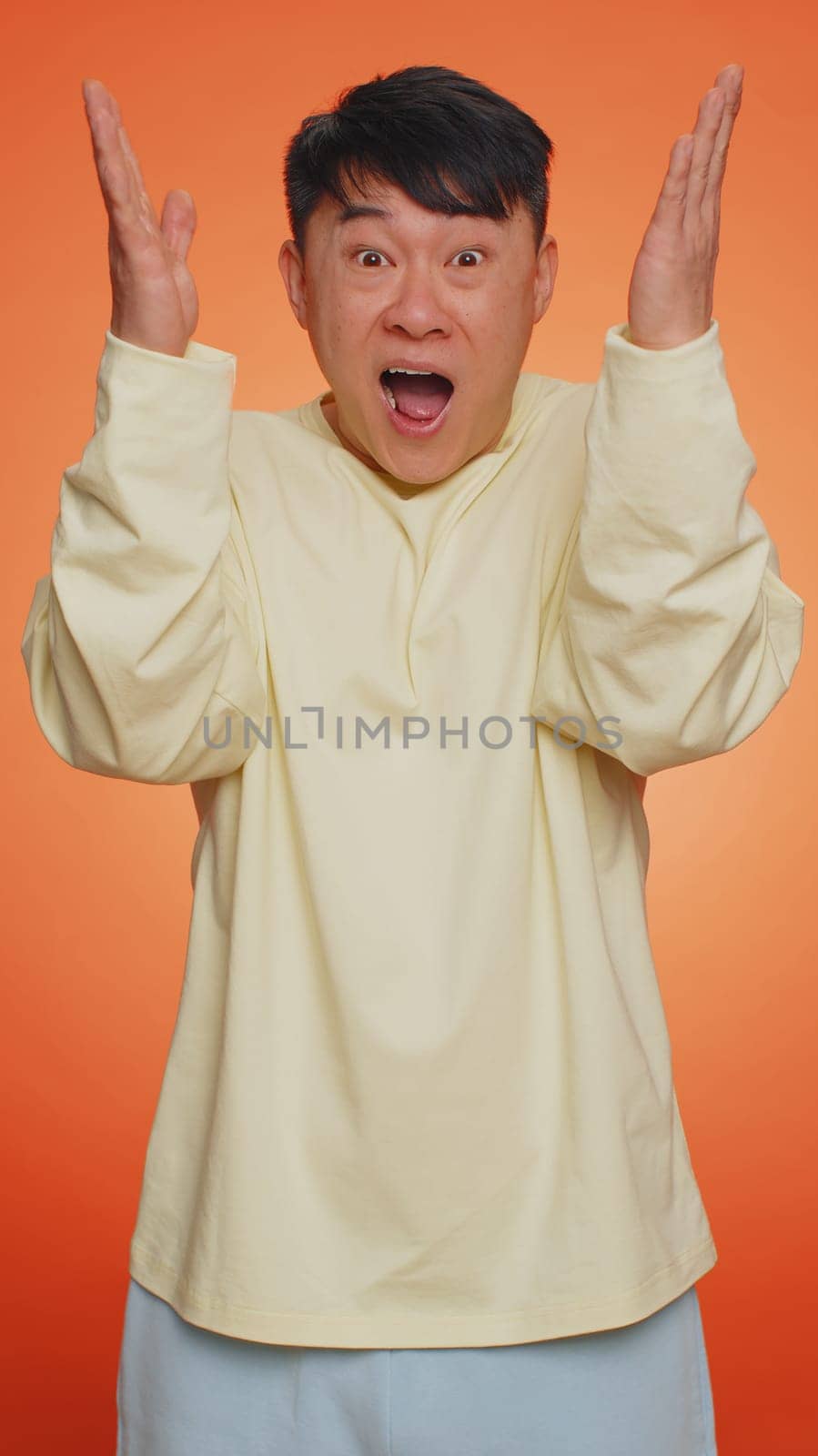 Excited amazed asian man touching head and showing explosion, looking worried and shocked, celebrating success idea. Looking surprised of win wow chinese guy on orange studio background. Vertical shot