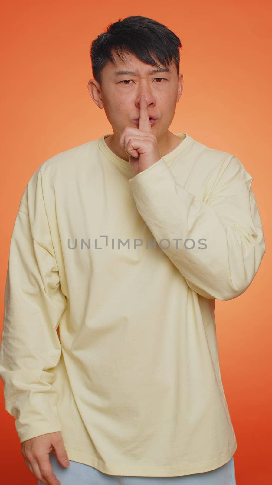 Keep my secret. Do not tell anyone. Shh be quiet please. Asian man presses index finger to lips makes silence gesture sign. Guy on orange studio background. People lifestyles. Vertical shot