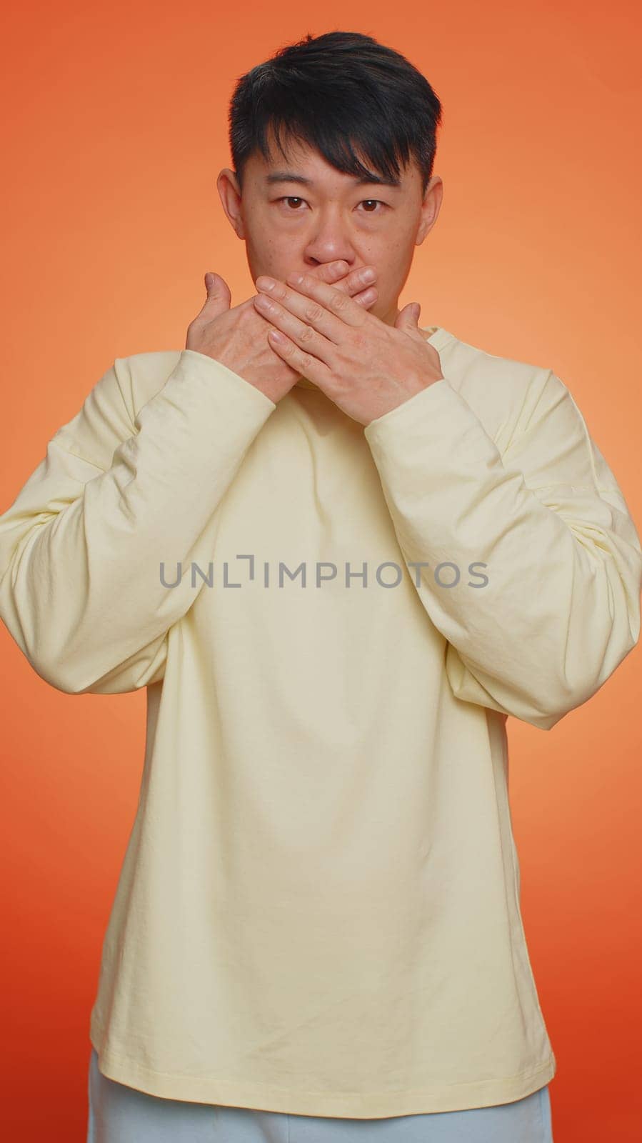 I will not say anyone. Frightened asian man closing mouth with hands, looking intimidated scared at camera. Guy gestures no, refusing to tell terrible secret, unbelievable truth. Vertical shot
