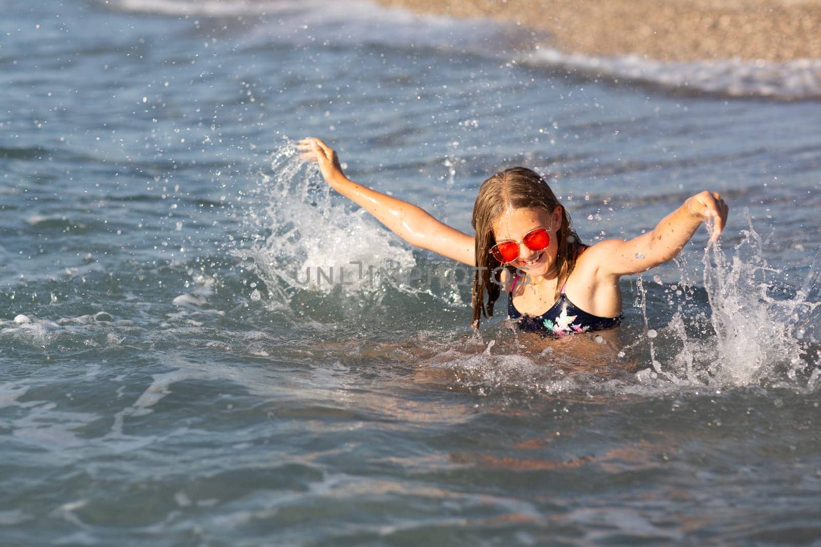 Teenage girl in pink sunglasses having fun on the beach in the sea, playing with waves and splashes. Fun on summer hloiday concept.