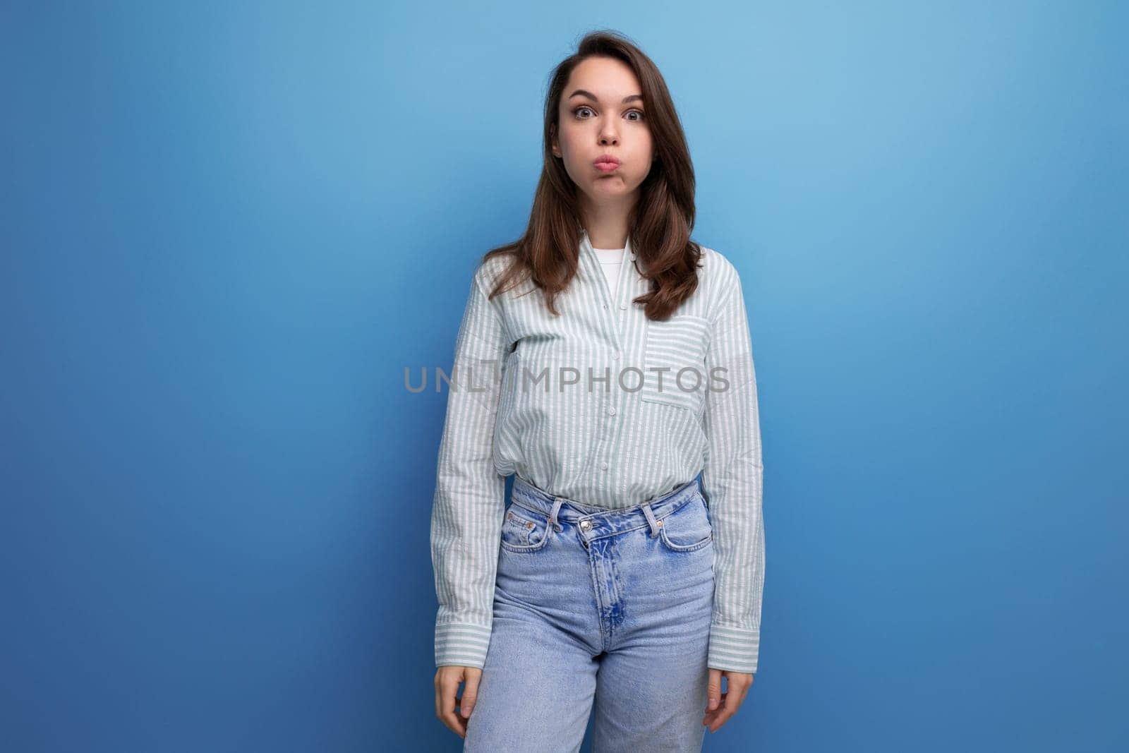 funny young brunette lady dressed in a striped shirt and jeans grimaces and makes a face.