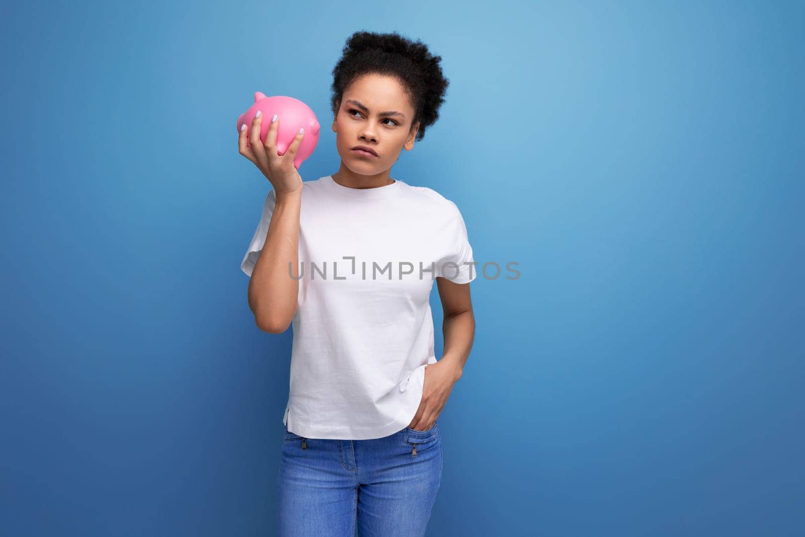 young pretty hispanic business woman in a white t-shirt holding a pink piggy bank on a blue background with copy space.