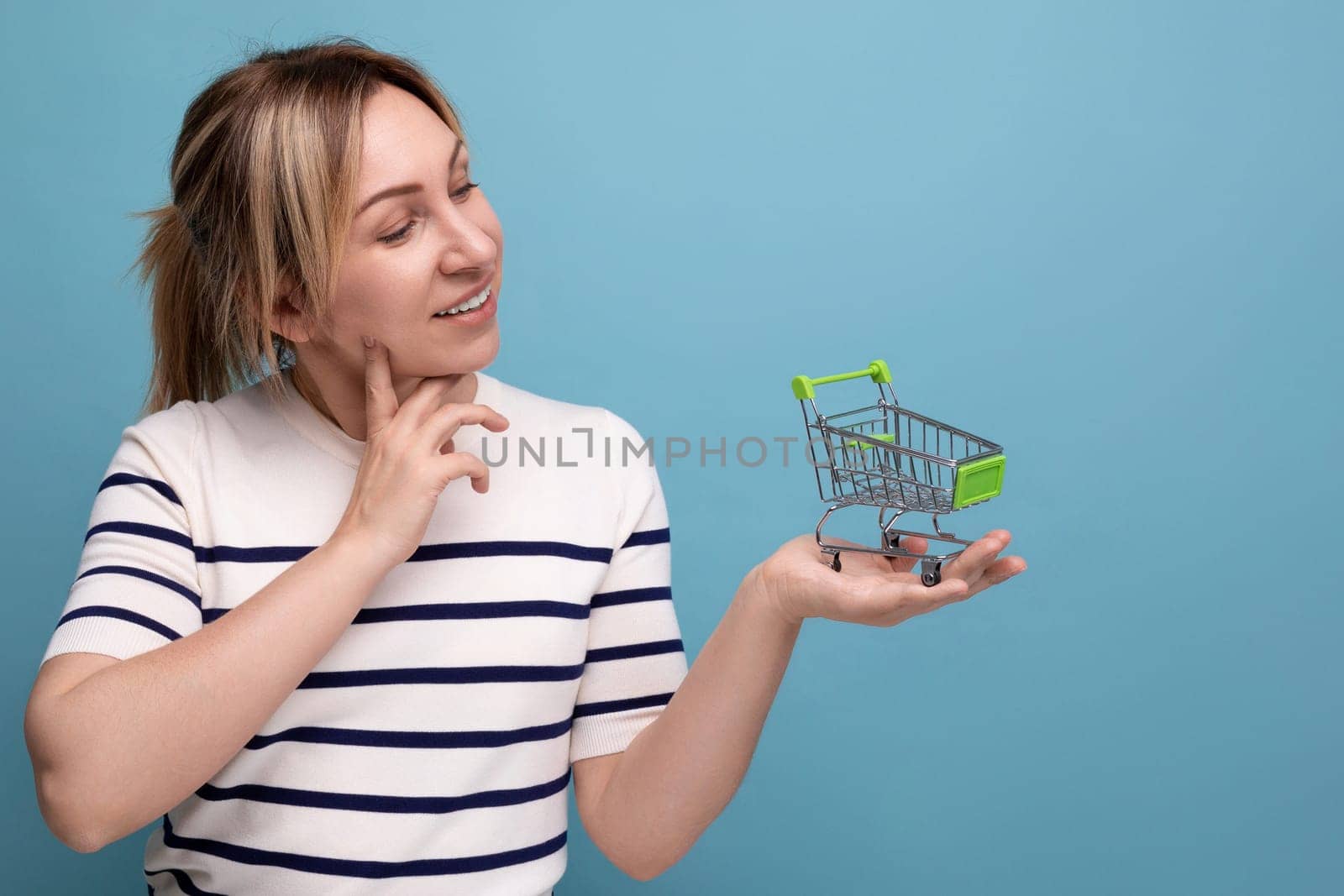 cute blond young mom shopaholic holding shopping cart and thinking how to spend money on blue background with copy space.