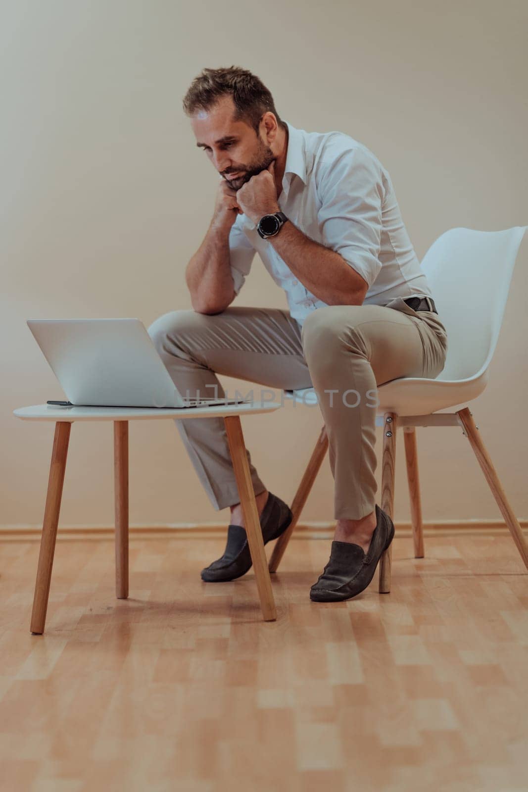A confident businessman sitting and using laptop with a determined expression, while a beige background enhances the professional atmosphere, showcasing his productivity and expertise