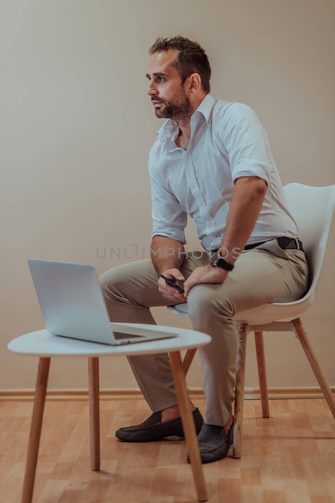 A confident businessman sitting and using laptop with a determined expression, while a beige background enhances the professional atmosphere, showcasing his productivity and expertise